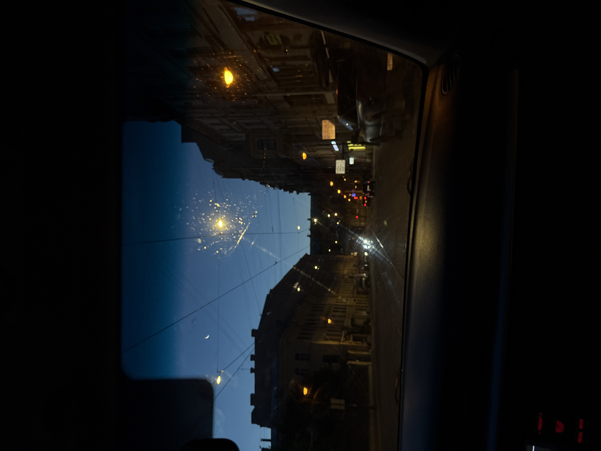 The image shows a view from inside a car looking through the windshield at a street scene at dusk. The sky is a deep blue, with a sliver of a crescent moon visible. Several streetlights are illuminated, casting a warm glow on the buildings lining the street. The buildings appear to be old, with multi-story facades and arched windows. A few cars are visible on the street, some moving and some parked. There are also overhead wires, likely for trolleybuses or streetcars, crisscrossing the sky. The windshield has some smudges and streaks, which are illuminated by the streetlights. The interior of the car is dark, with a dashboard visible at the bottom of the frame, including some illuminated buttons. The scene is likely a street in the historic center of Saint Petersburg, Russia, given the architectural style and the presence of trolleybus wires.