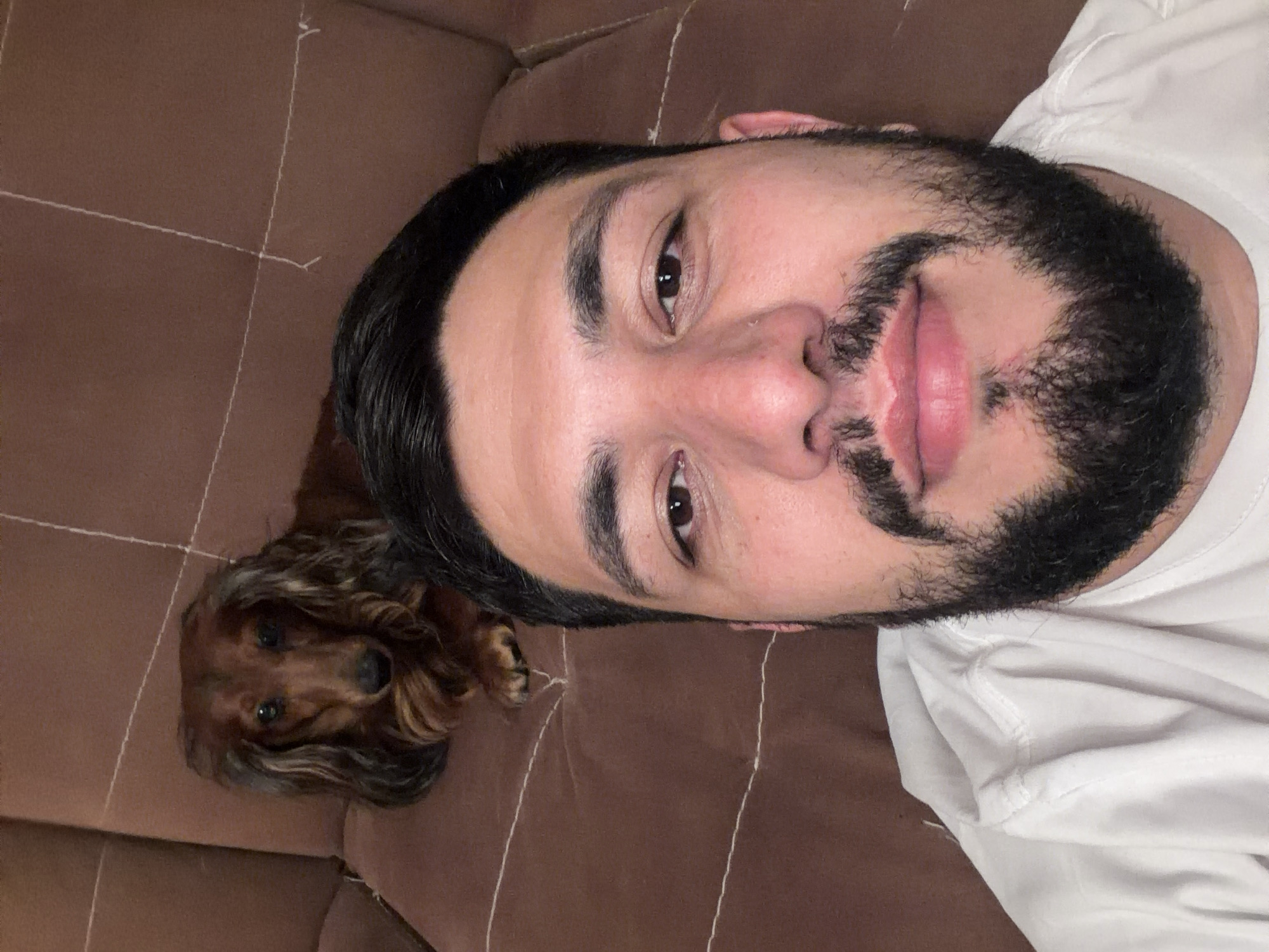 A man with dark hair and a beard is lying on a brown couch, next to a long-haired dachshund. The man is wearing a white t-shirt. The couch has vertical seam lines. The location is not identifiable as Krasnoyarsk, Russia based on visual information.