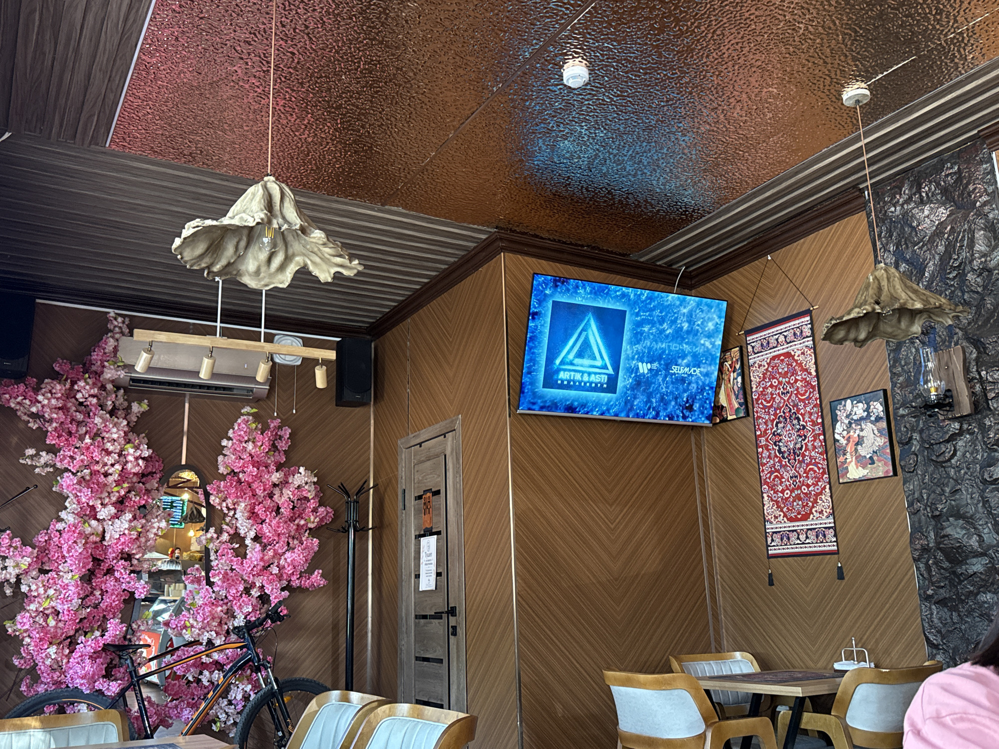The image depicts the interior of a cafe or restaurant, possibly located in Baikonur, Kazakhstan. The space features a unique ceiling with textured metallic panels in reddish-brown and dark brown hues. Two distinct, large, unconventional pendant light fixtures, resembling abstract, dried leaves, hang from the ceiling.
The walls are primarily covered in diagonally paneled wood, with a section on the right featuring a textured, dark gray, rock-like material. A prominent flat-screen television displays a vibrant blue abstract image with text overlay, likely advertising or a visualizer.
Decorations include a long, narrow red rug with intricate patterns hanging vertically on the wall, and a framed artwork with figures to its right. To the left, a large, artificial cherry blossom arrangement in vibrant pink dominates the space, framing a mirror that reflects a counter area with food displays and a person wearing a face mask. A bicycle is parked in front of the floral display.
Several tables with chairs are visible, some with cushioned seats and wooden frames, set in the foreground and midground. A coat rack stands near the cherry blossom arrangement. A door with a sign is located on the left wall. The lighting appears to be a mix of ambient and focused light, casting shadows and highlighting the textures of the space.