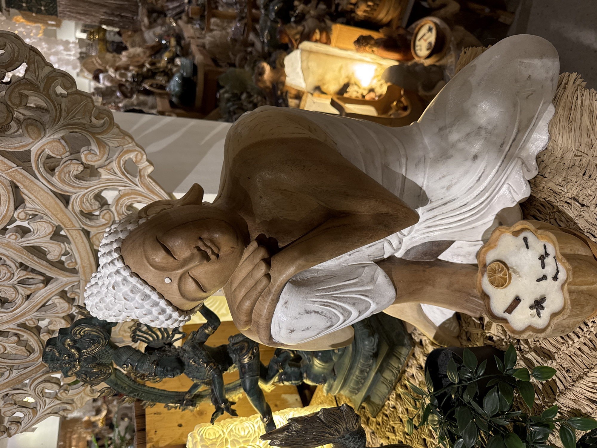 The image features a reclining Buddha statue. The statue is predominantly wood, with sections, such as the head covering and a portion of the robe, painted white. The Buddha is depicted lying on his side with his head resting on his hand. The background includes ornate, carved wooden designs and various decorative objects. In the foreground, there are plants and a candle adorned with spices and a dried citrus slice. The location is possibly in a shop or a temple. The location is Unknown, Indonesia.