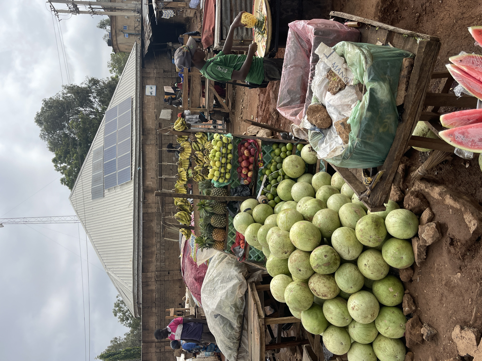The image depicts a vibrant outdoor fruit market in Gura Top, Nigeria, under a cloudy sky. The foreground is dominated by a large pile of round, green watermelons stacked on a wooden cart. Adjacent to the watermelons, there are sliced watermelons wrapped in plastic wrap. Other fruits like bananas, pineapples, apples, and citrus fruits are displayed on wooden shelves. A man wearing a green shirt and a cap is preparing fruit. Another person in a pink shirt stands nearby. A structure with solar panels is visible in the background. Above the wooden carts, bags filled with other produce, some labeled "MANGO", can be seen. The ground around the fruit stand is earthen, and there are rocks scattered around. Power lines are also visible above.