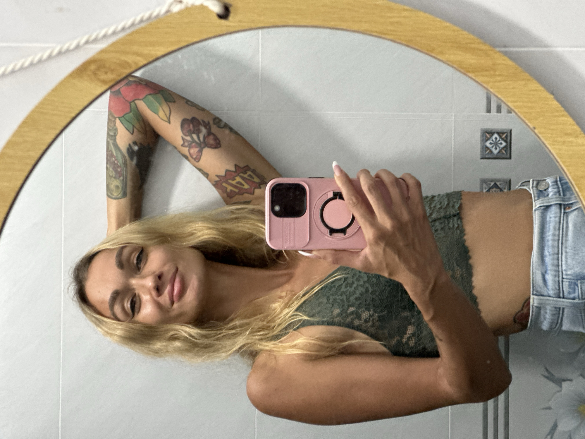 A woman with blonde hair, wearing a green lace bralette and blue jeans, takes a selfie in a bathroom mirror. Her left arm is raised, with tattoos visible on her upper arm. She holds a pink phone in her right hand. The bathroom has white tiled walls and a circular mirror with a wooden frame. The image is a medium shot, focused on the woman and her reflection. The scene appears to be indoors, likely in a private residence or hotel room. The background details are limited to the bathroom's tiled surfaces and a portion of the mirror's edge. The image does not contain any specific visual cues that would allow for the identification of its location in Mũi Né, Vietnam.