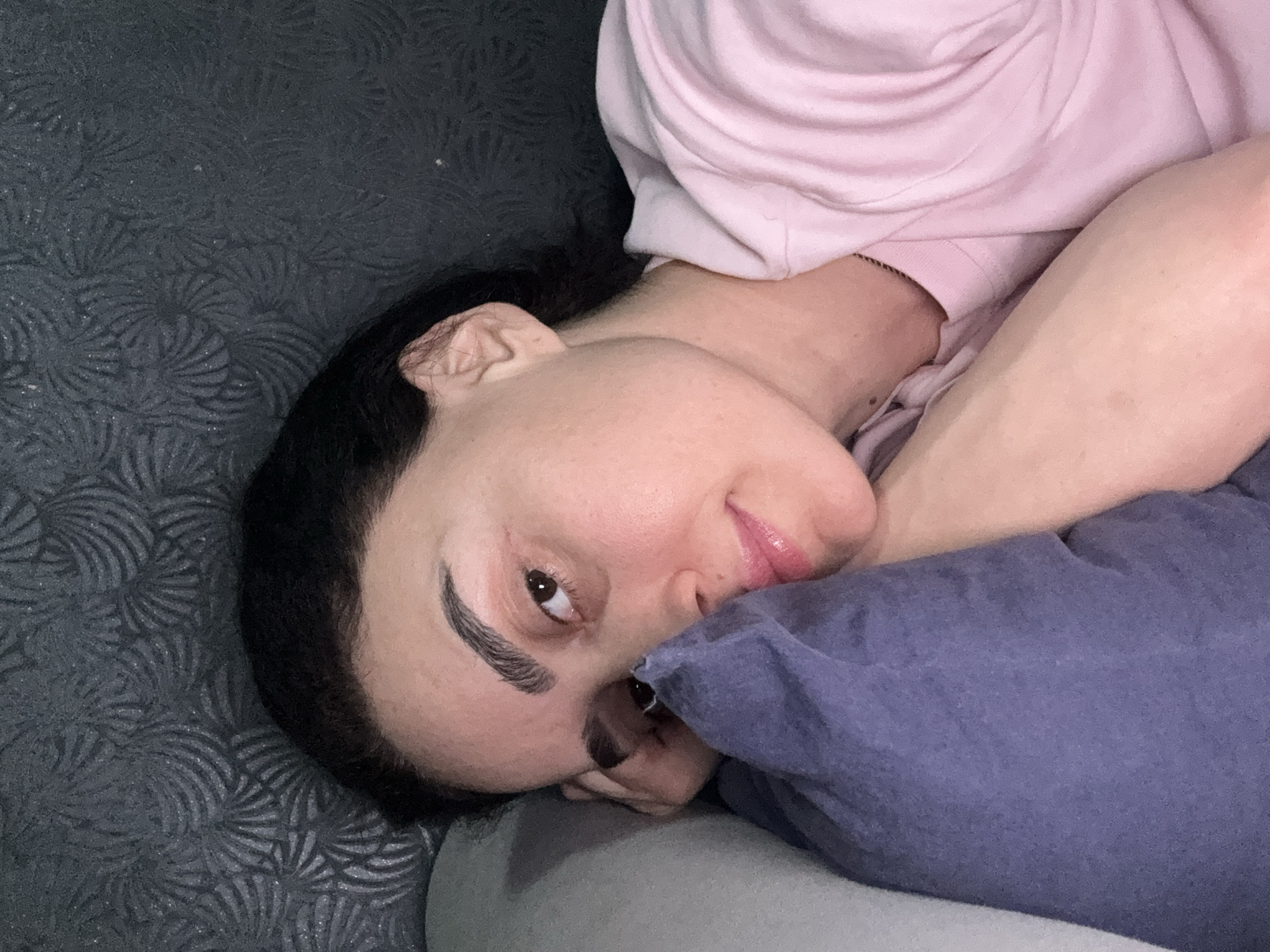 A woman is reclining on a grey surface with a patterned texture. She is resting on her right side with her head turned towards the camera. She is wearing a light pink top. Her left arm is crossed over her body and is partially covered by a blue pillow. Her hair is black, and her right eye is open and directed at the camera. The environment is indoors. The location in the city of Tbilisi, Georgia cannot be determined from this image.