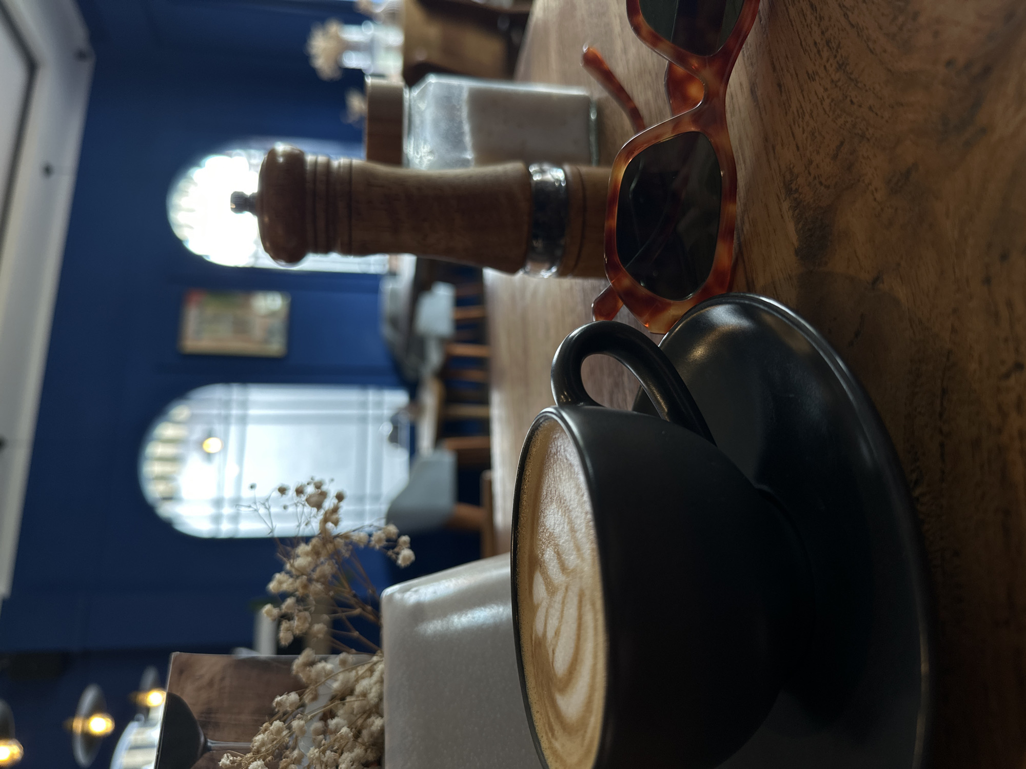 The image presents a focused, intimate tabletop scene within a cafe or restaurant, located in North Kuta, Indonesia. The main subjects are a dark ceramic coffee cup, filled with a creamy latte featuring visible latte art, resting on a matching saucer. Beside it, a pair of stylish tortoiseshell-patterned sunglasses with dark lenses is casually placed. These items are arranged on a warm, textured wooden table with a visible grain.
In the background, slightly out of focus, stands a tall, slender wooden pepper or salt grinder, alongside a clear glass container filled with white salt. To the left, a decorative element composed of dried, light-colored flowers in a simple white holder adds a subtle touch.
The setting features a deep blue wall in the distant background, punctuated by two distinct, arch-topped windows that allow abundant natural light into the space. Through one window, a blurred bright light, possibly a pendant lamp, is visible. The scene suggests a tranquil, daytime environment, conducive to a coffee break or a moment of relaxation. No people are visible in the frame, and no discernible text can be identified on any objects or surfaces. The overall ambiance is calm and inviting.