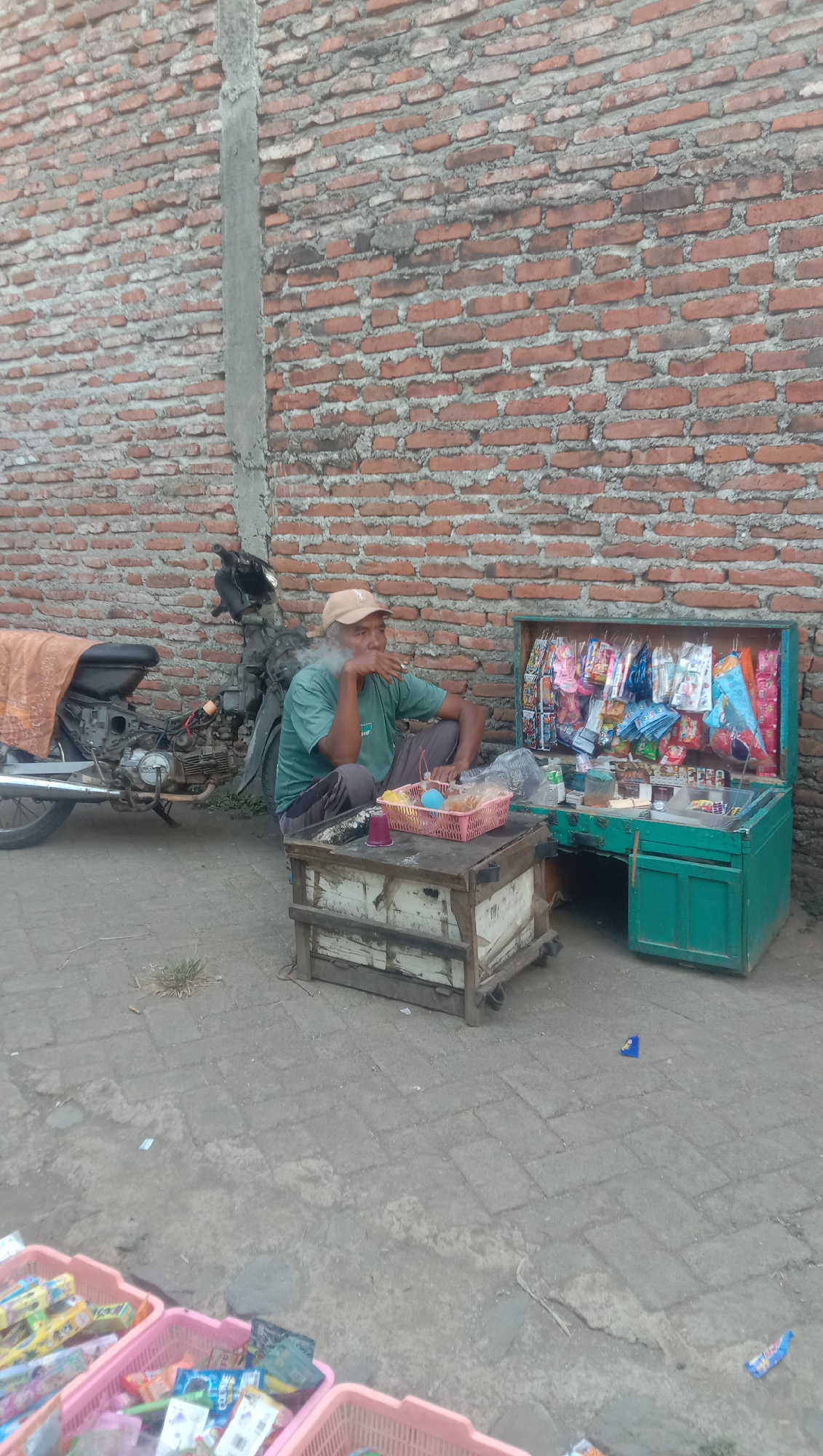 An outdoor scene in Karang Anyar, Indonesia, depicts a man sitting on the ground next to a roadside stall. He is wearing a green t-shirt, dark pants, and a beige cap, and is smoking a cigarette. The stall, painted teal, displays various packaged snacks and candies. In front of the stall, a wooden cart with wheels holds a pink basket filled with snacks and two cups. A motorcycle, covered with an orange cloth, is parked behind the man. The background consists of a red brick wall with a concrete pillar. The ground is paved with interlocking gray bricks. In the foreground, three pink plastic baskets are filled with merchandise.