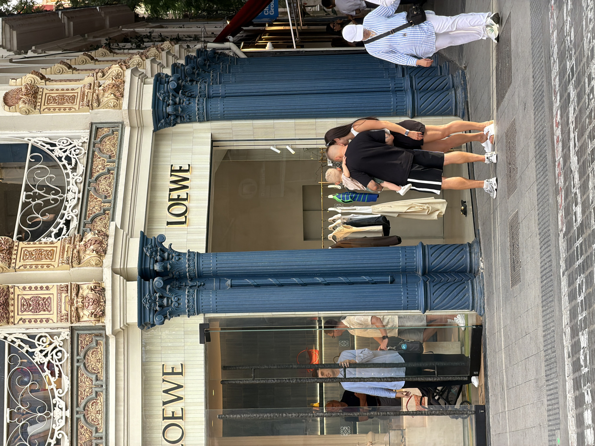 In the city of Unknown, Unknown, a Loewe store is depicted. The building's facade features ornate, decorative elements and blue fluted columns. In front of the store, a group of people are visible. A man in a light blue shirt and black pants stands inside the store behind a glass partition, looking towards the right. Another man in a white patterned shirt and grey shorts is to his right, also within the store. Outside the store, a couple stands with a young child being held by the man. The man wears a black t-shirt and black shorts with white stripes, while the woman is in a black top and dark shorts. Further to the right, a person in a blue and white striped shirt and white pants walks away from the camera. The street in front of the store is paved with cobblestones and has visible metal grates. Racks of clothing are displayed outside the store entrance.