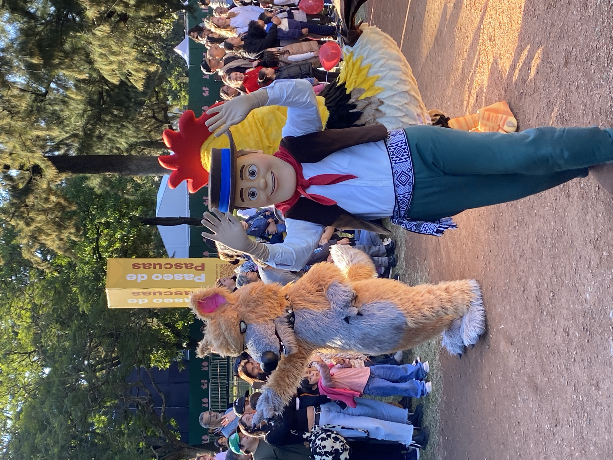 The image captures a scene in what appears to be a public park setting. The location is in Buenos Aires, Argentina, as evidenced by a sign in the background. In the foreground are two costumed characters. One is dressed as a fox and is surrounded by a crowd of people, including a young child in a pink shirt. The other character is dressed as a man with what appears to be a rooster design on the back. There are numerous other people in the background, standing and watching. The environment suggests a sunny day in a park or recreational area with trees and open space. The background includes a mix of adults and children. The overall scene is festive, indicative of an event or public gathering.