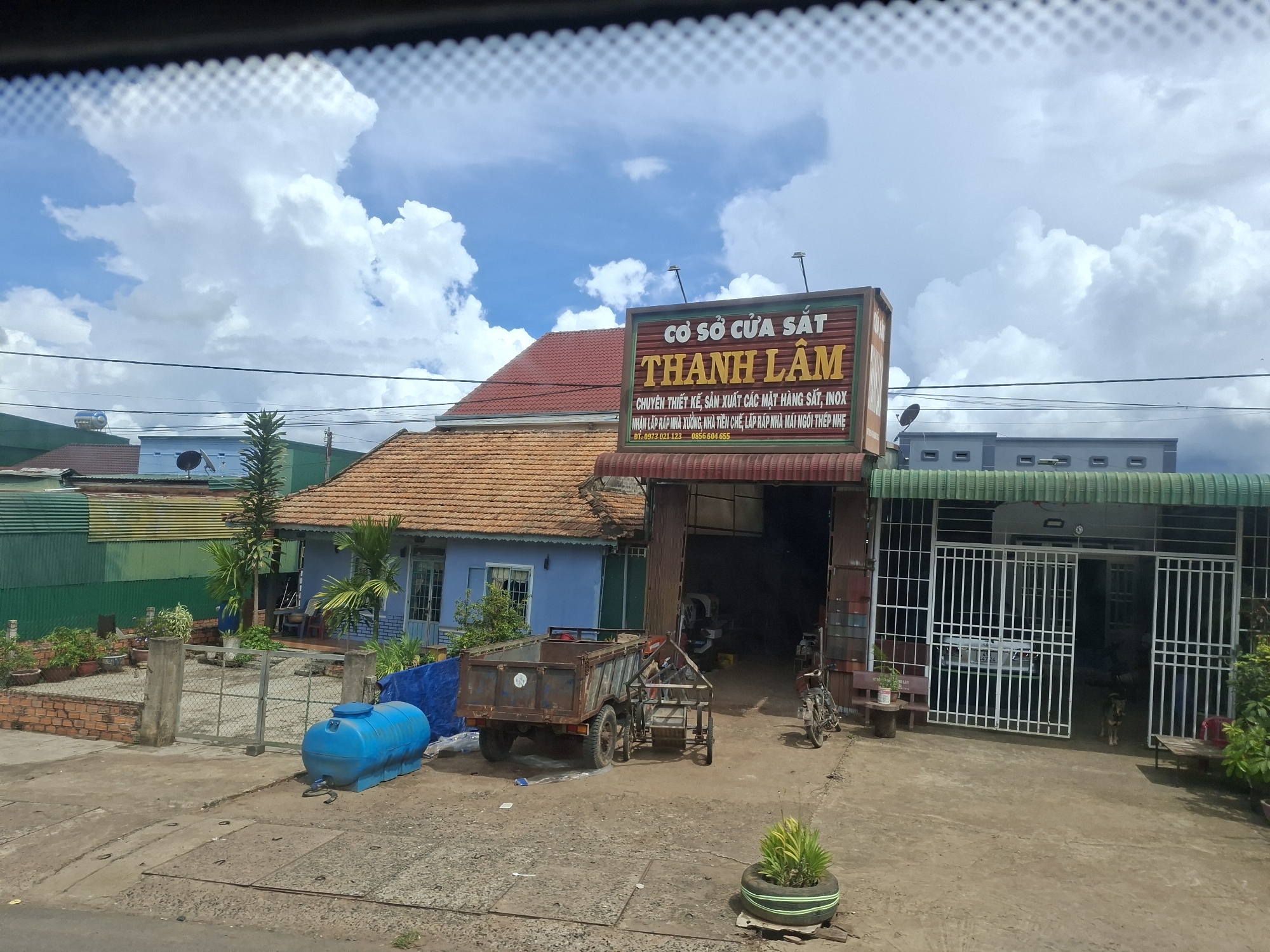 The image shows a street view of a building with a sign that reads "CƠ SỞ CỬA SẮT THÀNH LÂM" in Vietnamese, indicating a metal gate and door workshop. The building is of moderate size with an orange tiled roof and a blue-painted facade. In front of the building, there is a workshop area with a rusty truck and a small trailer. A blue water tank is located on the left side of the scene. A white metal fence with a gate is visible in front of a property next to the workshop. The area appears to be a commercial or industrial zone. The sky is partly cloudy.