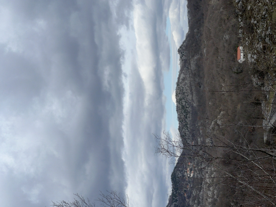 The image displays a rural mountainous landscape in Vrela, Montenegro, under an overcast sky. Approximately two-thirds of the frame is filled with layered dark grey and white stratocumulus clouds, with isolated patches of light blue sky visible at the horizon. In the foreground and mid-ground, several bare deciduous trees with slender, dark branches are present, with some branches extending into the frame from the left. The terrain consists of exposed grey rock formations, dry grasses, and sparse brown vegetation. In the background, undulating hills and mountains are extensively covered with leafless deciduous trees and rocky outcroppings. On the upper left background hillside, a small cluster of buildings with orange-red roofs constitutes a discernible settlement. On the lower right background hillside, a solitary, white-walled house with a prominent orange-red tiled roof is situated amidst rocky and sparsely vegetated terrain. No individuals or specific actions are discernible.