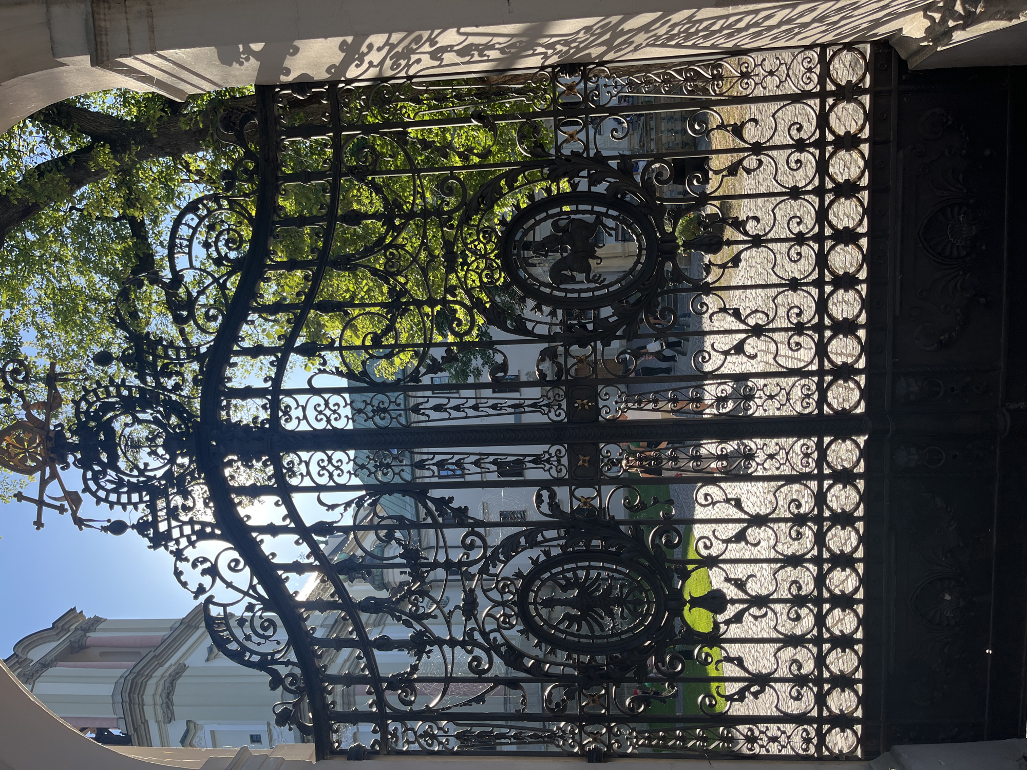 The image shows a large, ornate wrought-iron gate in front of a building, likely in Krakow, Poland, given the architectural style and heraldic motifs. The gate is elaborately decorated with scrolling metalwork, circular medallions, and finials. One medallion prominently displays an eagle, likely representing Polish heraldry, while another features a rider on a horse. Sunlight streams through the gate, casting shadows on the cobblestone ground and highlighting the intricate details of the ironwork. Behind the gate, parts of a light-colored building with classical architectural elements are visible, along with lush green trees. The sky is clear and blue. A few blurred figures of people can be seen in the background, suggesting the location is accessible to the public. The overall impression is one of historical grandeur and artistic craftsmanship.