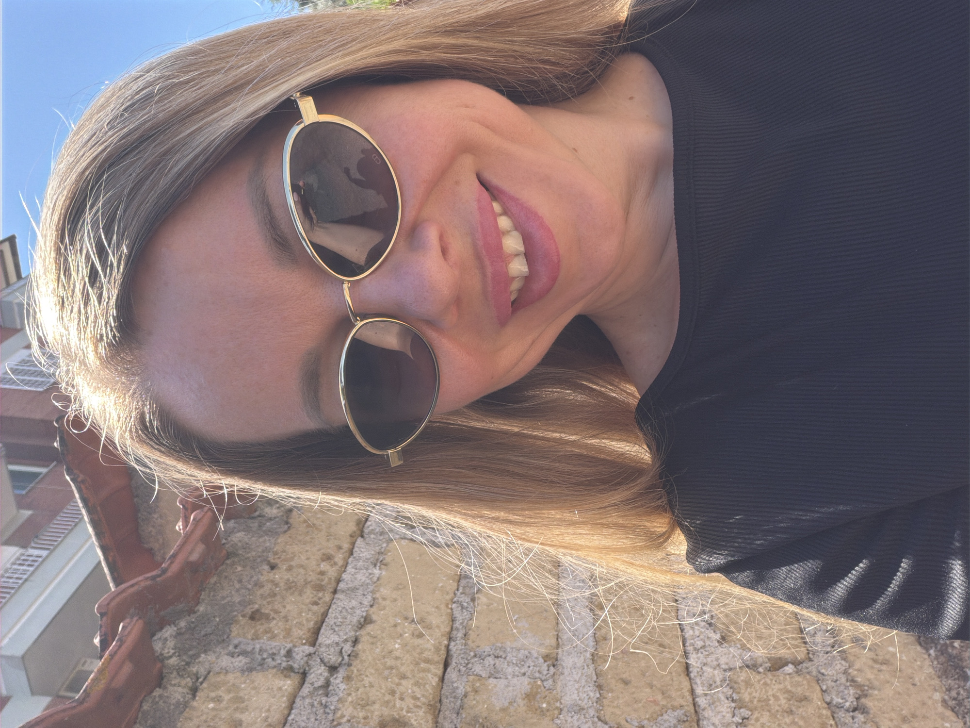 A close-up selfie shows a woman with shoulder-length blonde hair wearing gold-rimmed oval sunglasses and a black ribbed top. She is smiling and her lips are painted pink. She stands in front of a yellow brick wall. The sunlight casts shadows on her face and hair. A portion of a red tile roof and a building with balconies are visible in the background. The scene appears to be outdoors on a sunny day. The specific location in Rome, Italy, is not identifiable from the image.