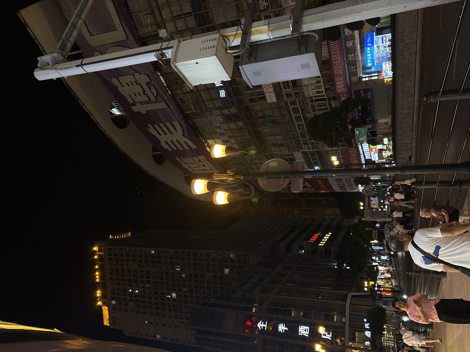 The image depicts a street scene at night in Yuzhong District, China. Streetlights illuminate the area, revealing tall buildings and shops with illuminated signs in Chinese characters. People are seen walking and standing on the sidewalk, including individuals wearing casual clothing. A large sign with Chinese characters is visible above the street. The area appears to be a commercial district, with various businesses and advertisements present.