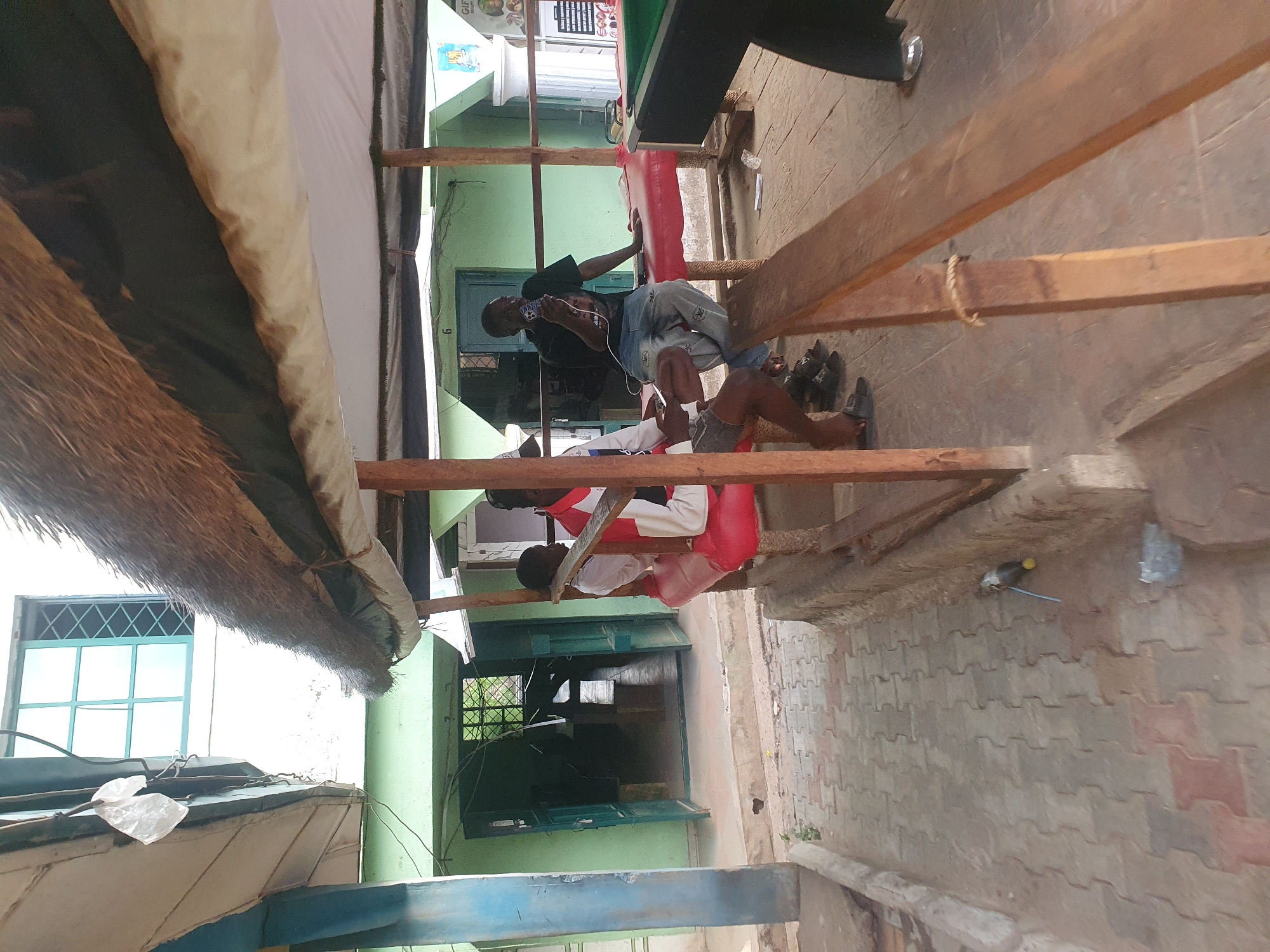 This media file captures an outdoor or semi-outdoor scene, possibly a casual gathering area or part of a market in Lokoja, Nigeria, during the daytime.
In the foreground and midground, several men are present. One man, wearing a black t-shirt and light-colored shorts or jeans, is seated or leaning against a red cushioned bench. He is holding a phone to his ear, seemingly engaged in a conversation, with white earphones or a wire dangling from the phone. Another man, partially visible to his left, wears a red and white jacket. Further to the left, only the legs and feet of a third man, wearing shorts and dark shoes, are visible. The men appear relaxed.
The setting features a makeshift shelter or canopy constructed from wooden beams and poles. Part of the roof is covered with light-colored fabric, while another section is made of woven straw or thatch. In the background, light green-painted buildings are visible, some with windows featuring blue frames and grid patterns, and others with dark security grills. A pool table with a green surface is partially visible at the upper right.
The ground is paved with grey and reddish interlocking bricks. Scattered on the ground are several pairs of dark shoes and a plastic bottle with a yellow cap and blue straw. Some white plastic bags are also seen on what appears to be a blue railing or structure in the lower left. In the far background, a blurry sign reads "GIFT BEAUTY," with additional unreadable text below it. The overall impression is of a casual, everyday scene.
