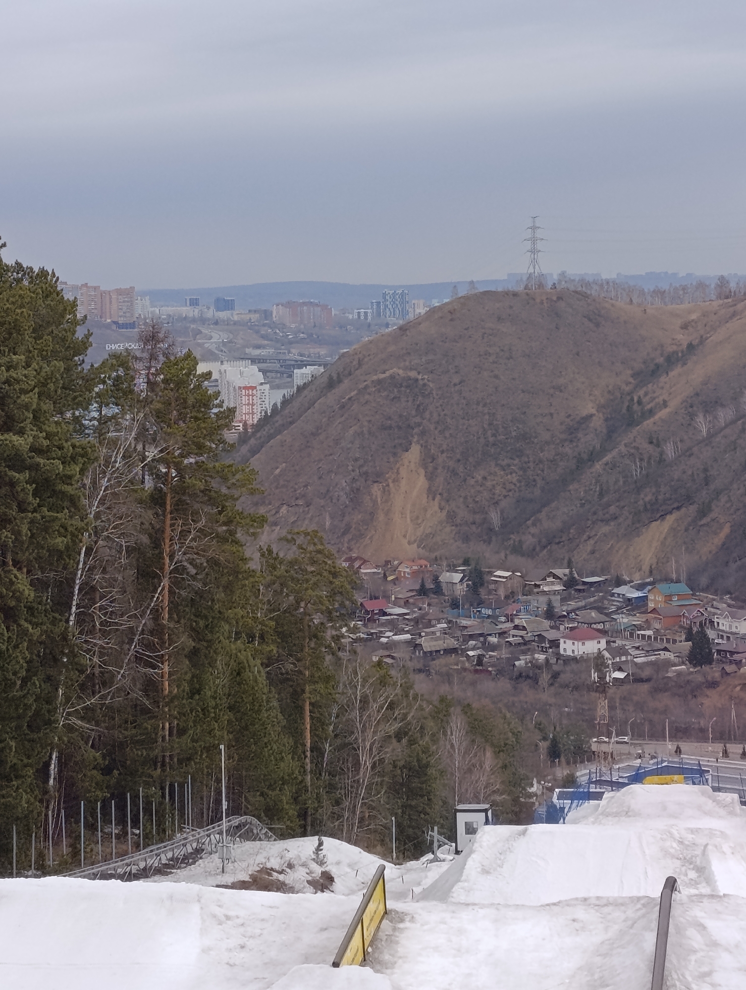 The image depicts a view of Krasnoyarsk, Russia, from what appears to be a ski or snowboarding park overlooking the city. In the foreground, there is a snow-covered ramp and ski run with some metal fencing. Mid-ground shows a valley containing a cluster of houses and buildings, indicative of a residential area. The background includes a range of hills, with one prominent hill sporting an electrical transmission tower on its peak. Further in the distance, the urban landscape of Krasnoyarsk is visible, with buildings and structures forming the city skyline under an overcast sky. The presence of snow suggests it is winter or early spring.