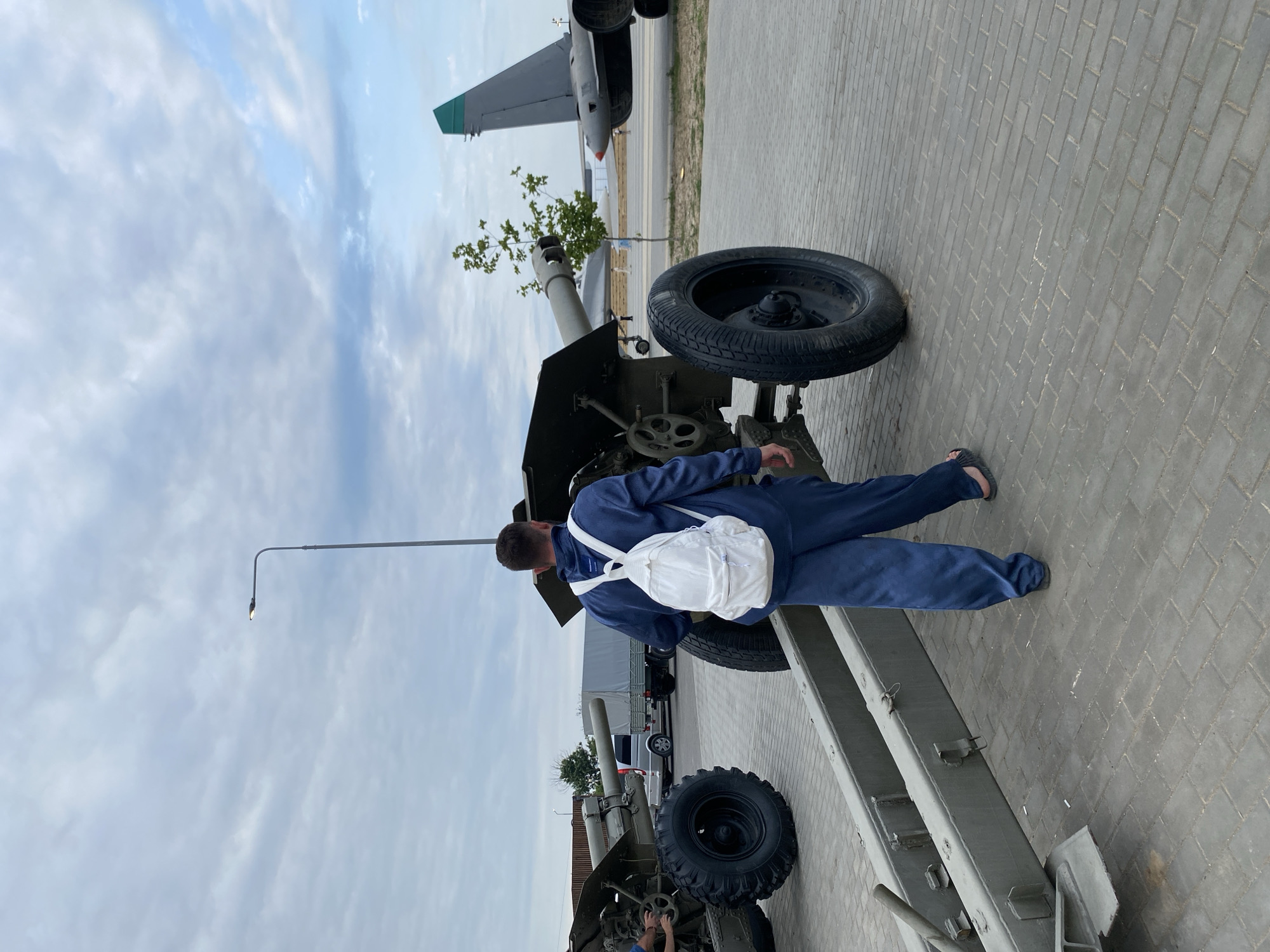 In Arablyar, Russia, a person wearing a dark blue jumpsuit and a white backpack walks away from the camera on a paved area. The person is positioned between two large, green artillery pieces. The artillery pieces are of military design, with large wheels and cannons. In the background, a gray truck with a tarped cargo bed is visible, as are two aircraft, one with a distinctive green tail fin. The sky is overcast with scattered clouds.