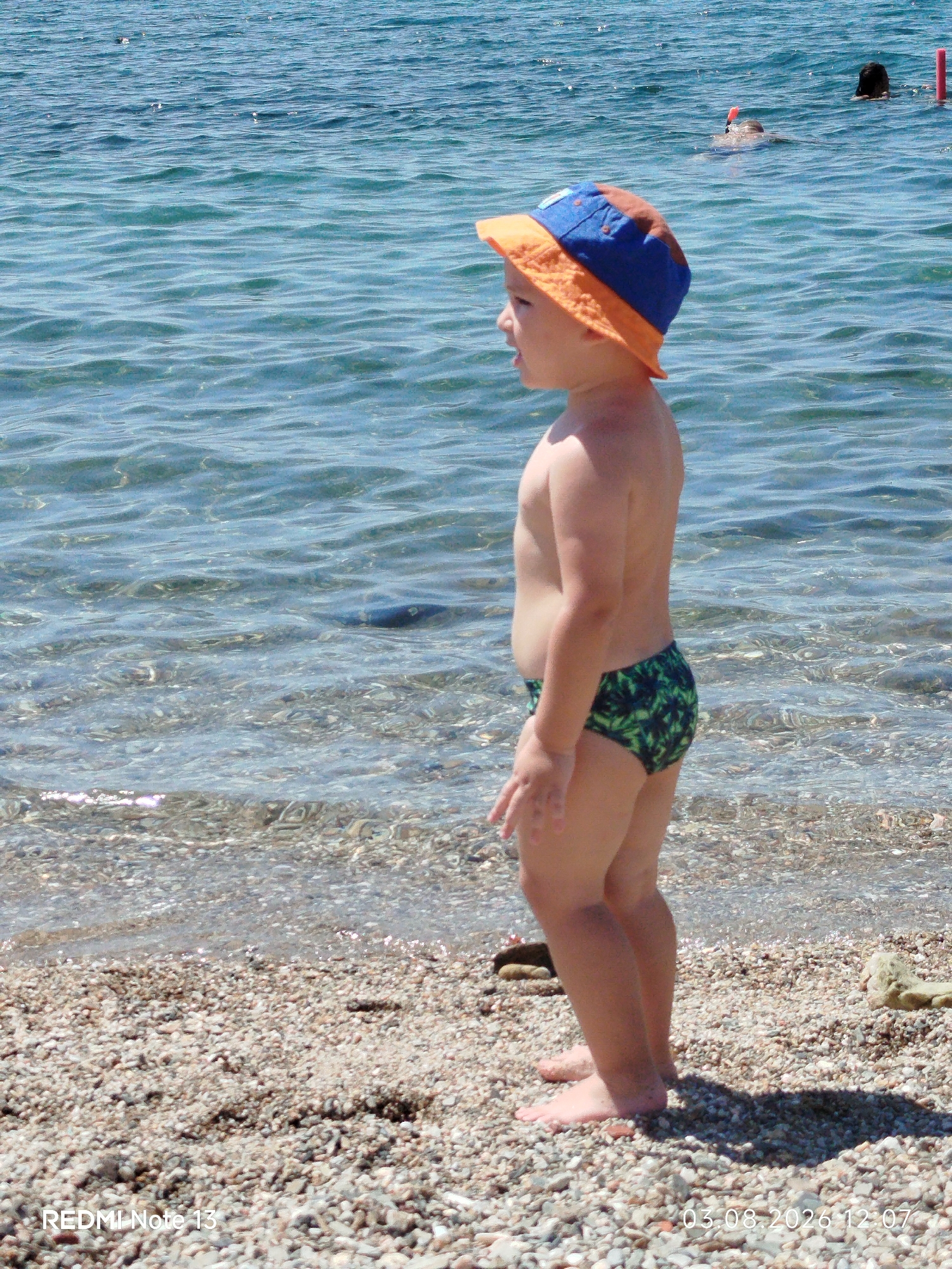 The image depicts a child standing on a pebble beach at the sea in Sithonia, Greece. The child is wearing a blue and orange sun hat and green and black patterned swim trunks. The child is facing left, looking out at the water. The sea is calm and clear, with small waves. In the background, two other people are visible swimming in the sea. The sun is shining, casting shadows on the beach. A timestamp is visible in the bottom right corner, reading "03.08.2026 12:07". The text "REDMI Note 13" is visible in the bottom left corner.