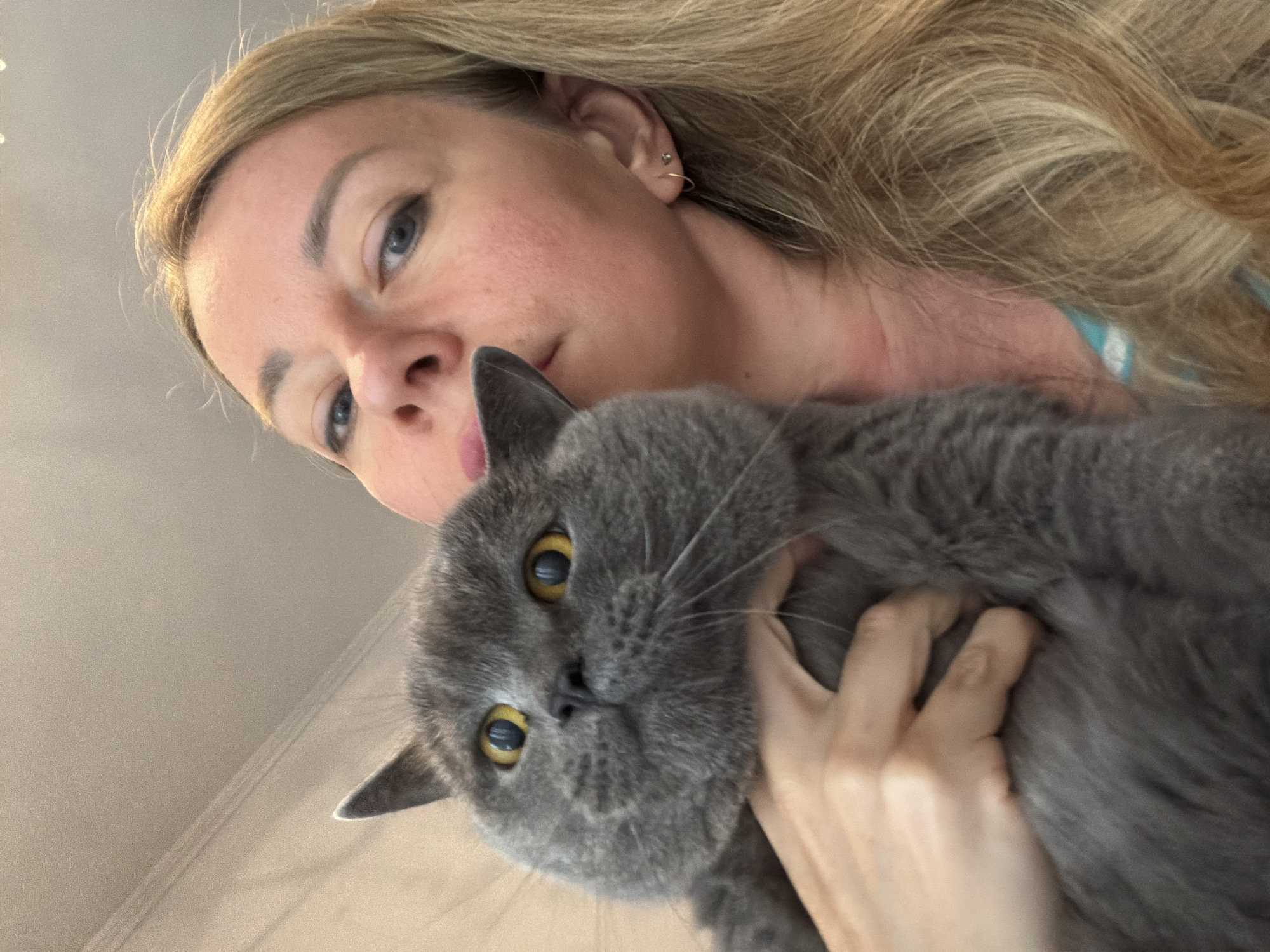 A close-up selfie shows a woman holding a grey cat. The woman has blonde hair and is looking at the camera. The cat has yellow eyes and is looking away from the camera. The background is a beige wall. The scene is indoors and appears to be in a domestic setting. There is no indication of the city of Rome, Italy in the image.