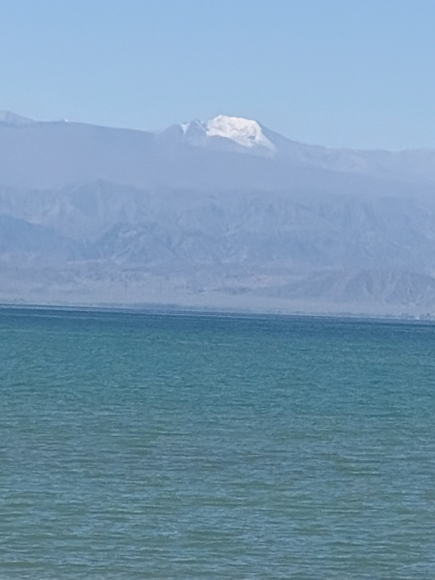 The image shows a vast body of calm, turquoise water in the foreground. In the middle ground, a range of hazy, blue mountains stretches across the horizon. The distant peaks of these mountains are capped with white snow, indicating a high altitude. The sky above is a clear, pale blue. The scene depicts a natural landscape with water and mountains under a bright sky.