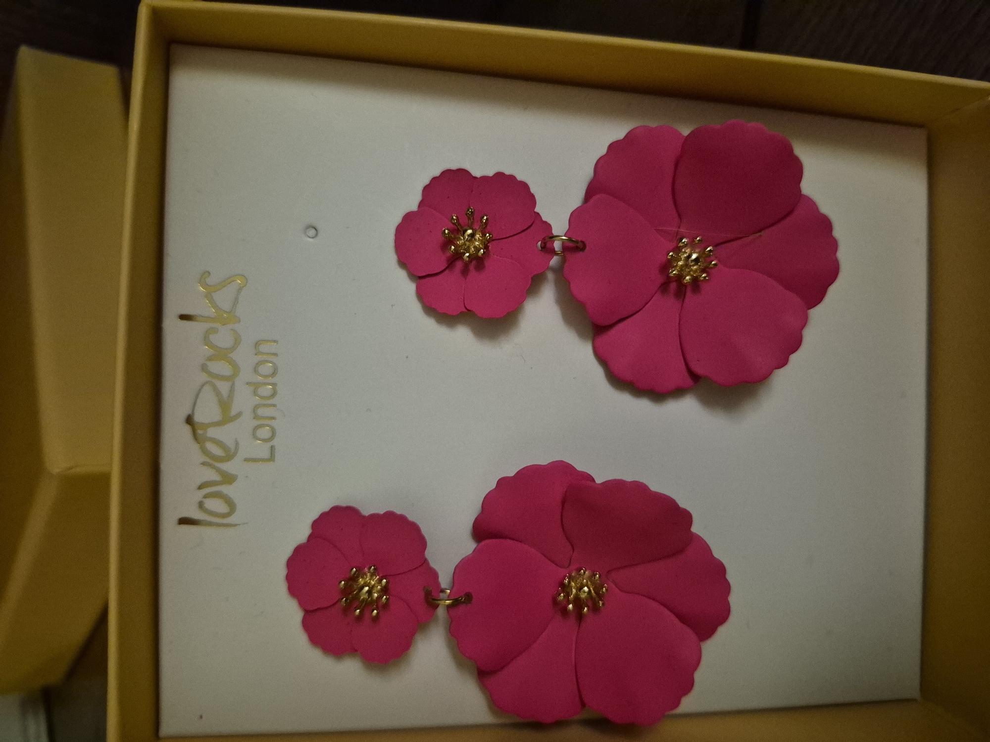A pair of dangling floral earrings is displayed in a yellow box with the "loveRocks London" logo printed in gold. The earrings are a bright fuchsia color and are shaped like flowers with gold centers. Each earring has two layers of petals, with the larger flower hanging below a smaller one. The box is open, and the earrings are placed on a white backing. The background of the image is blurred and appears to be a dark surface.