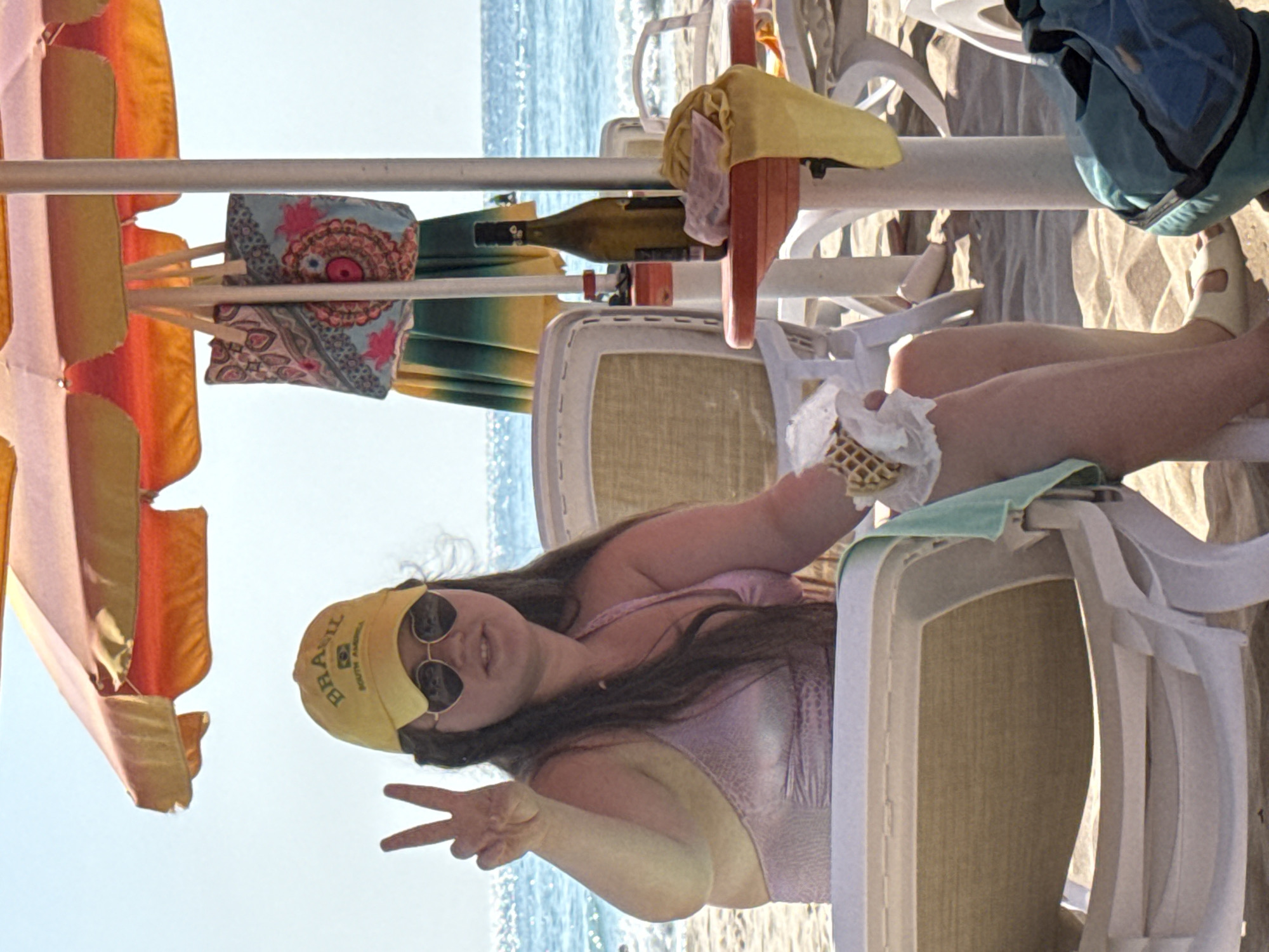 A woman wearing a pink bikini and a yellow baseball cap with "BRASIL" written on it is sitting on a beach chair under an orange and yellow umbrella. She is holding a waffle cone wrapped in white paper and making a peace sign with her left hand. She is also wearing heart-shaped sunglasses. To her right, on a small red table, is a bottle of wine and a yellow towel. The scene is set on a sandy beach with the ocean in the background. This image was taken in Obrochishte, Bulgaria.