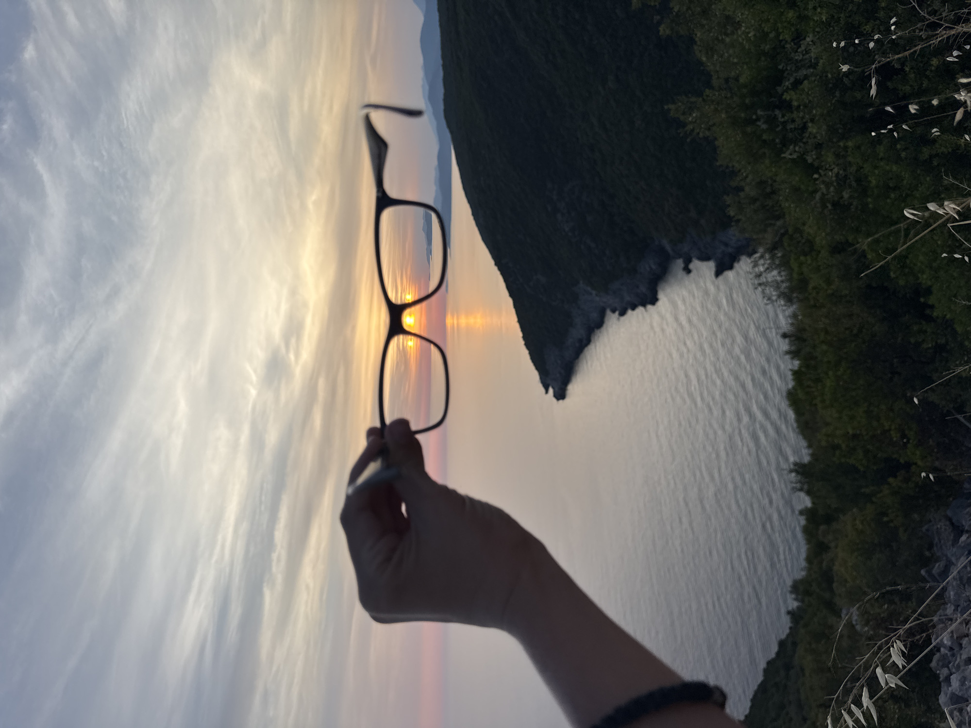 In Zagora, Montenegro, a person holds a pair of glasses in the foreground, framing a picturesque sunset over the Adriatic Sea. The sky is a canvas of soft pastel hues, with wispy clouds catching the dying light of the sun, which is positioned just above the horizon. Through the lenses of the glasses, the sun appears as two distinct, glowing orbs, casting a shimmering reflection on the calm water. The coastline, covered in lush green vegetation, curves into the distance, leading the eye towards hazy, blue mountains on the horizon. The sea is a deep, tranquil blue, with subtle ripples indicating a gentle breeze. The overall scene evokes a sense of peace and natural beauty.