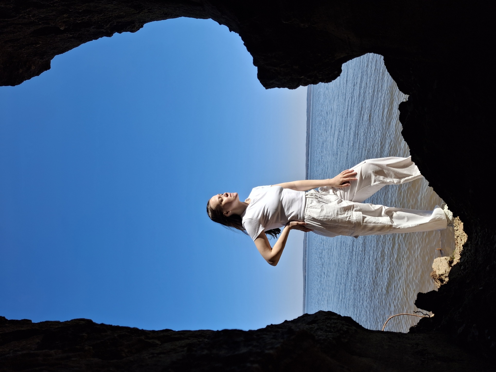 A wide-angle shot from inside a dark cave shows a woman standing near the edge. The cave entrance frames a bright blue sky above and a body of water on the horizon. The woman is wearing a white t-shirt and pants and stands with one hand on her hip. The lighting is bright outside the cave, contrasting with the darkness inside. The location in the city of Unknown, Unknown.