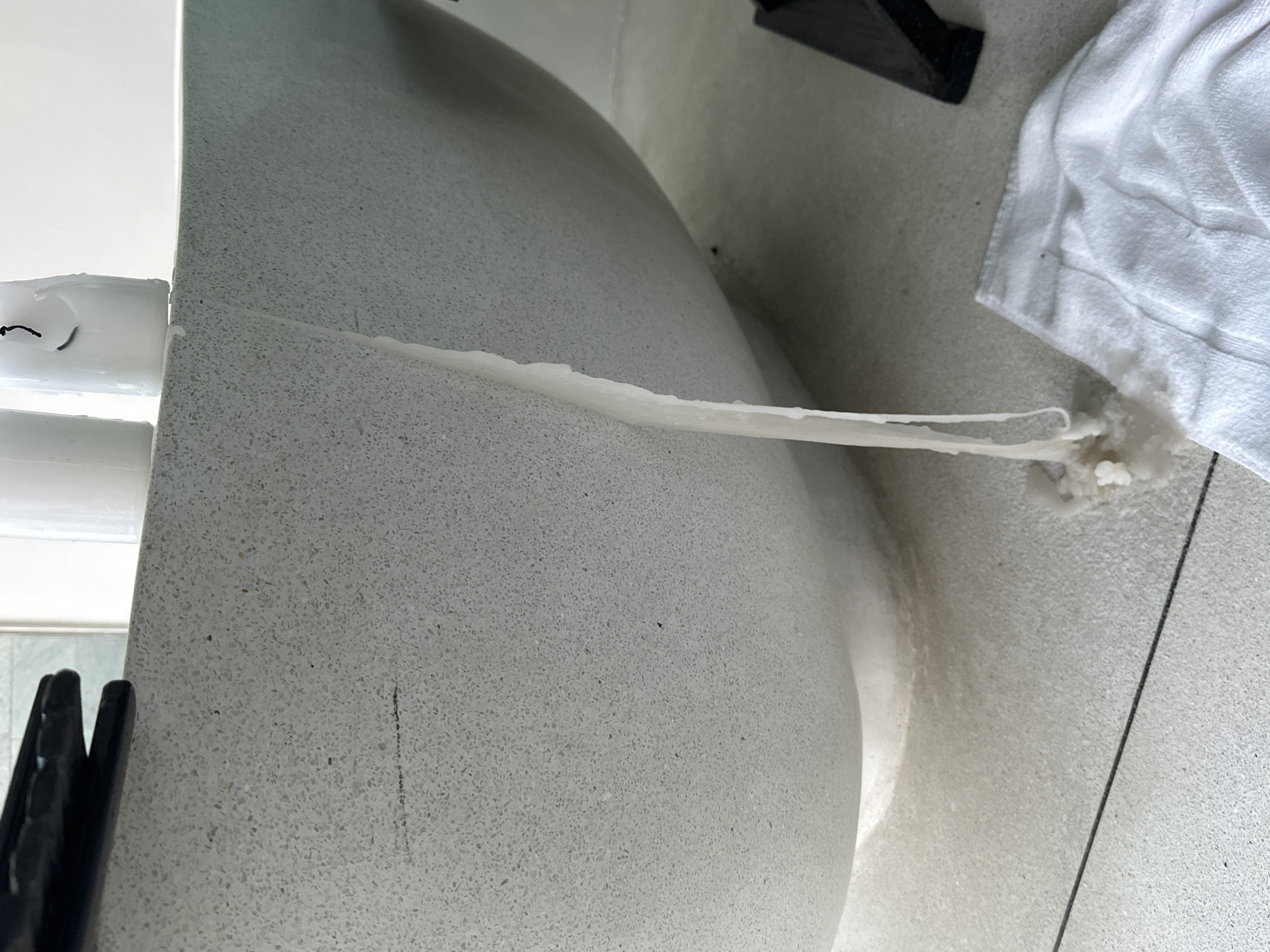 In Ko Samui District, Thailand, a close-up shot reveals a contemporary, speckled grey bathtub with a thick stream of white wax dripping from its rim down its side. The wax has pooled on the floor below, with a white towel placed nearby. The overall scene suggests a recent event involving lit candles near the bathtub.