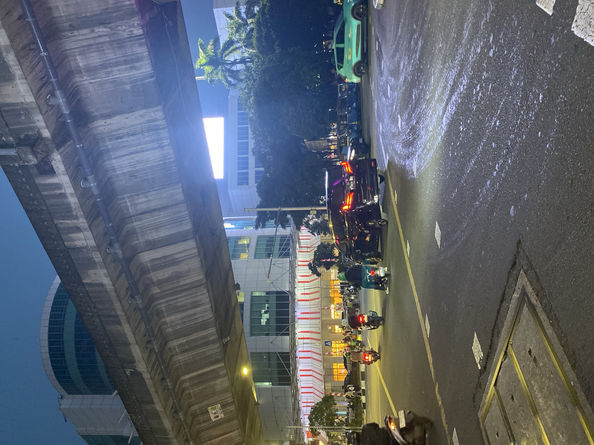 This image captures a nighttime scene on a street in South Jakarta, Indonesia, likely near a commercial or transportation hub, under an elevated structure. In the foreground, a dark-colored minivan with its brake lights illuminated is turning right on a wet asphalt road, with streaks of white residue visible on the surface, suggesting recent rain or some spilled substance. Several motorcycles are also on the road, with riders in casual attire. Some are in motion, while others appear stationary.
In the middle ground, a multi-story building with a curved facade and large windows is visible. Below the building's upper floors, a section is adorned with alternating red and white striped awnings, suggesting a commercial establishment. A few pedestrians are seen near the entrance of this building. Palm trees are interspersed between the structures, indicating a tropical urban environment.
The upper portion of the image is dominated by the underside of a large, concrete elevated structure, possibly a highway or a train line, showing signs of wear and staining. Pipes and conduits are attached to its underside. The sky is dark, indicating it is night, with a bright rectangular light panel visible in the distance, possibly a large digital advertisement or a screen. A turquoise colored car is visible in the far right of the scene, also on the street.
