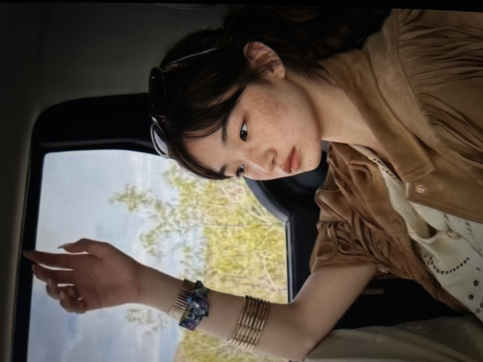 A young East Asian woman with freckles is sitting in a car, looking out the window. Her right arm is raised, with her hand resting on the car's window frame. She is wearing a brown fringed jacket over a white embroidered top. She has several bracelets on her wrist, including a stack of gold bangles and a patterned fabric bracelet. Sunglasses are resting on top of her head. The background outside the window appears to be a dry, grassy landscape with scattered trees under a cloudy sky. The interior of the car is dark.