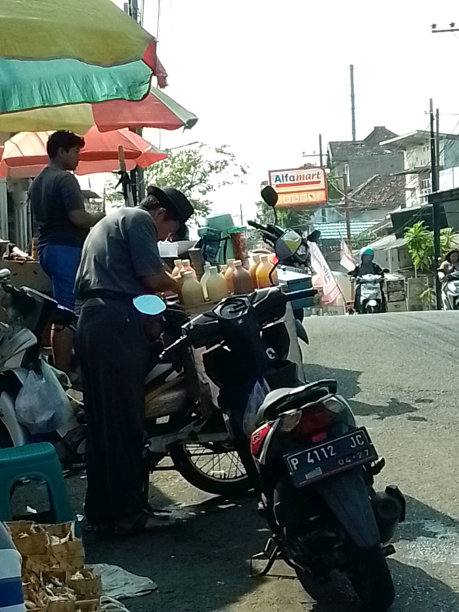 The image depicts a street scene in Jember, Indonesia, with a focus on two individuals interacting near a stall of bottled beverages and motorcycles. In the foreground, a person wearing a dark shirt and pants, with a hat, is bent over a table filled with bottles, possibly preparing or selling them. A younger person in a dark t-shirt and blue shorts stands nearby. Several motorcycles are parked around the stall, including one in the immediate foreground with a license plate reading "P 4112 JC 04-27". In the background, a sign for "Alfamart" is visible, indicating a commercial area. Other motorcycles are seen on the road, with riders, suggesting ongoing traffic. The environment appears to be an outdoor market or roadside vendor area, under awnings providing shade. The overall lighting suggests it is daytime, with clear sky.
