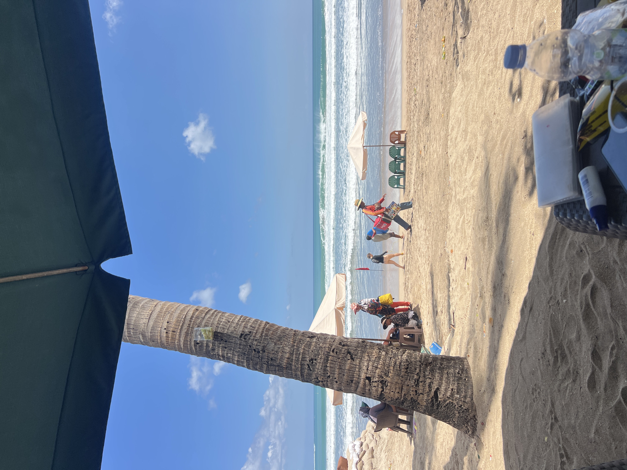 The image depicts a sunny day at Kuta Beach in Kuta, Indonesia. In the foreground, a large palm tree trunk dominates the left side of the frame, with a dark green beach umbrella casting a shadow over the sandy ground. In the mid-ground, people are scattered on the beach. To the left, two individuals sit in chairs near a sandbag barrier. Closer to the water, a small group gathers under a beige umbrella, with one person in a colorful outfit holding a yellow bag. Further to the right, a person wearing a straw hat walks across the sand, carrying a red bag and some papers. Several empty beach chairs are arranged in front of another umbrella. The ocean stretches to the horizon with moderate waves. The sky is a clear blue with a few white clouds. On a table in the lower right corner, a water bottle, a packet of tissues, and a tube of sunscreen are visible.