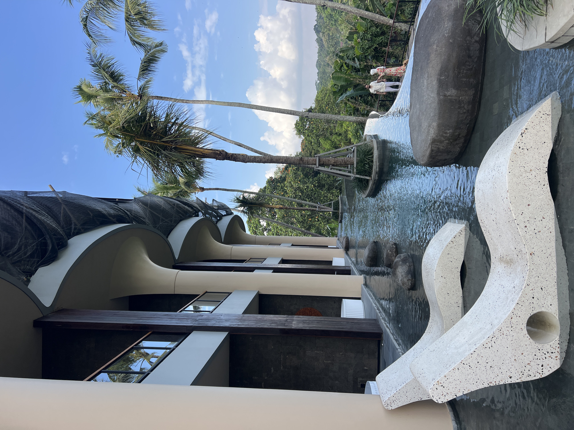 In the city of Ubud District, Indonesia, a serene infinity pool curves around a modern building with flowing architectural lines. The pool's edge is marked by several large, smooth rocks, and two white, speckled lounge chairs are partially submerged in the shallow water, their sleek design contrasting with the natural elements. Beyond the pool, a lush green landscape of tropical trees and foliage stretches out, with a clear blue sky dotted with fluffy white clouds overhead. Two individuals, a man in a light-colored shirt and pants and a woman in a floral dress and headscarf, stand on a wooden walkway overlooking the view, a small dog at their feet. The building's facade features large columns, dark wood accents, and balconies with glass railings, suggesting a resort or high-end accommodation. The overall atmosphere is one of tranquil luxury amidst a vibrant natural setting.