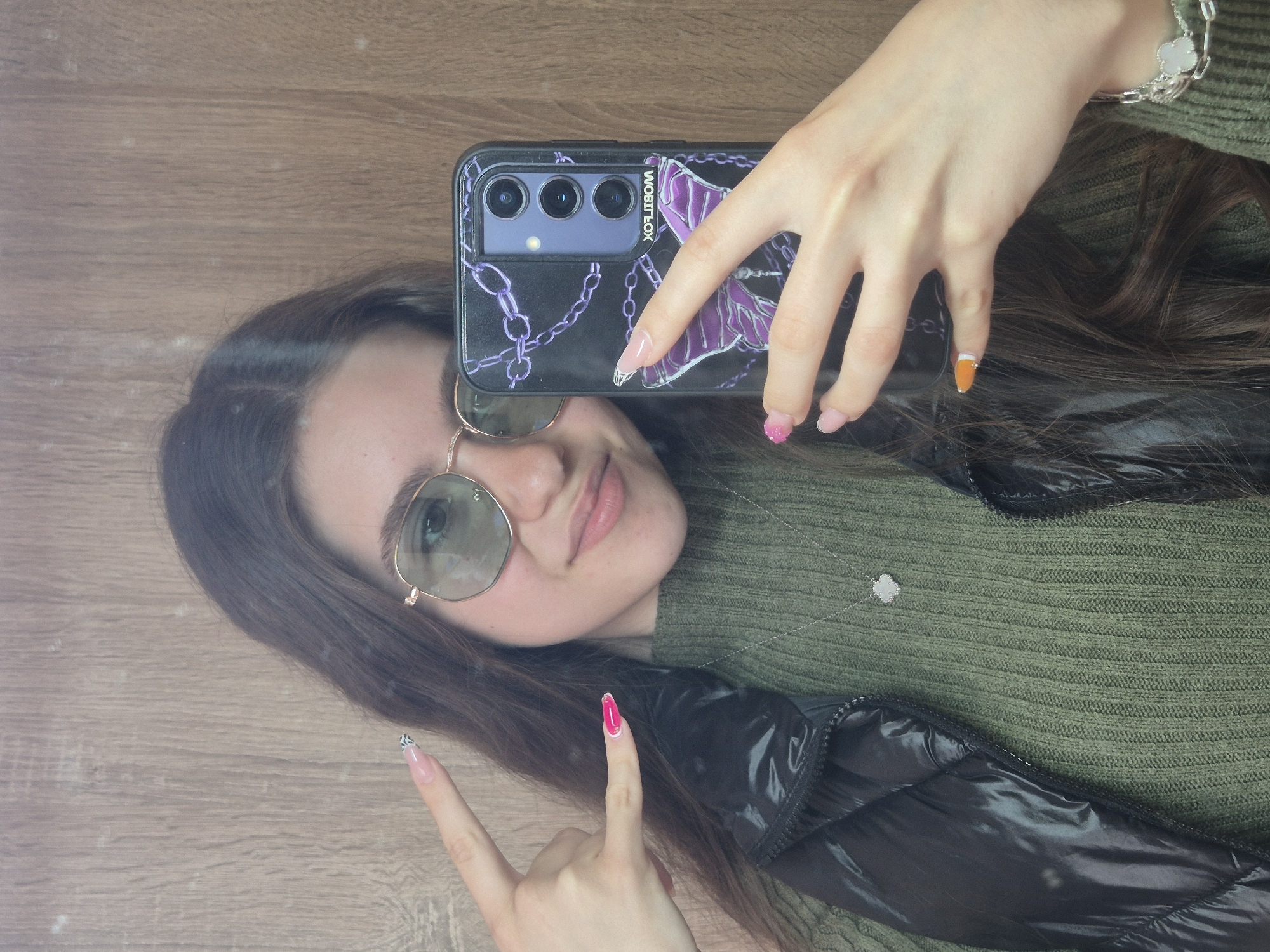 The image displays a young woman's reflection, captured as she takes a selfie. The reflection shows her lying horizontally against a light brown wooden surface. Her upper body is visible, dressed in a ribbed olive-green sweater and a black shiny puffer jacket. She has long brown hair and wears light-tinted, round metal-framed sunglasses. Her expression is a subtle smile.
In her right hand, she holds a black smartphone with a distinctive case featuring purple chains and a skeleton/butterfly design. The phone has three visible camera lenses, and the text "WOBITFLOX" is visible on the side of the case. Her right hand also sports a silver bracelet with a four-leaf clover charm. Her fingernails are long and almond-shaped, with various designs: the pinky is bright pink with glitter, and other nails have clear/light pink bases with white designs.
Her left hand is raised, forming a "rock on" gesture with the index and pinky fingers extended. The nails on this hand also feature diverse designs: the index finger has a black and white abstract pattern, the middle finger is bright pink, the ring finger has a clear base with a white design, and the pinky finger is bright orange with a white design. A delicate silver necklace with a small white charm, possibly another clover, is visible on her chest. The scene is set in Pavlikeni, Bulgaria.