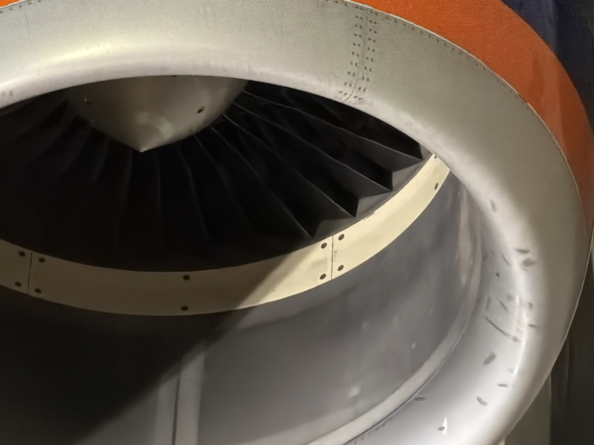 The image shows a close-up view of an airplane engine. The focus is on the fan blades within the engine, which are black and appear to be made of metal. The outer casing of the engine is silver and has a textured surface. There is an orange stripe running along the edge of the engine casing. The image is taken from a low angle, looking into the engine. The environment appears to be an airport or a hangar. There are no people visible in the image. The overall impression is one of mechanical precision and power. The location of Saint Petersburg, Russia, is not visually indicated in the image itself.