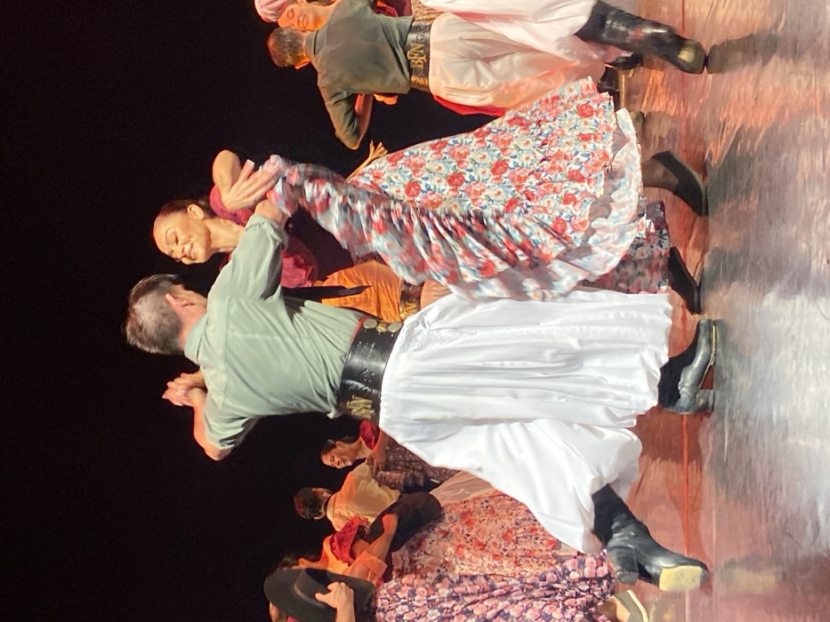 On a stage in Buenos Aires, Argentina, a group of dancers performs a folk dance. In the foreground, a male and female dancer are engaged in a partnered movement. The male dancer, with his back partially to the viewer, wears a sage green long-sleeved shirt, a dark neckerchief, a wide black leather belt featuring metal circular ornaments (*rastra*-style), wide white pants (*bombachas de campo*), and black leather boots with heels. His right arm is bent, and he is holding the female dancer's left hand. He has short, dark hair.
The female dancer, facing him and smiling, wears a maroon ruffled top and a flowing, multi-layered skirt with a red, blue, and white floral pattern. Her skirt shows motion blur, indicating dynamic movement. She wears dark, low-heeled shoes. Her dark hair is pulled back.
Behind the central pair, several other dancers are visible, performing similar movements. To the left, a dancer holds a dark, wide-brimmed hat. Other male dancers wear similar green or brown shirts and white pants with belts; one belt prominently displays "BEN". Female dancers in the background wear various floral patterned skirts. The stage floor is dark and reflective, illuminated by warm stage lighting that creates reflections. The background is unlit and dark, emphasizing the performers.