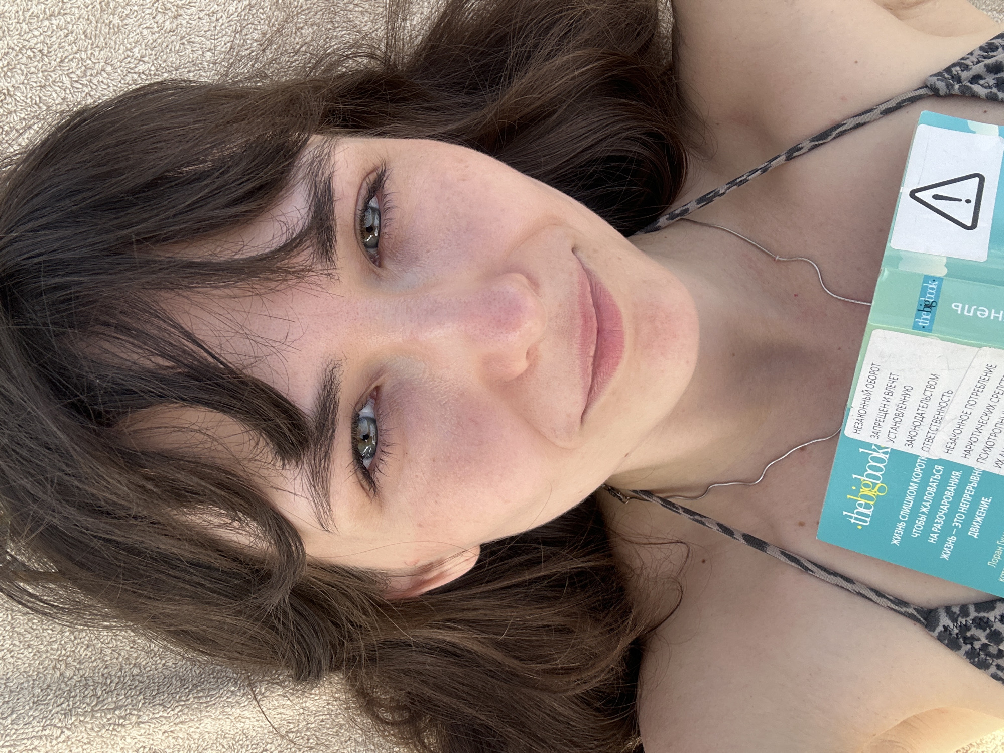 A woman with brown hair and green eyes is lying on a tan-colored surface, possibly a beach towel. She is wearing a bikini top with a leopard print. A silver necklace rests on her chest. In her right hand, she is holding a blue and white book titled "The Big Book", with text in Russian. The book has a warning sign on the cover. The photo has been taken from a high angle, looking down on the woman. The location is not explicitly mentioned, and there's no obvious evidence indicating it is Alanya, Turkey.