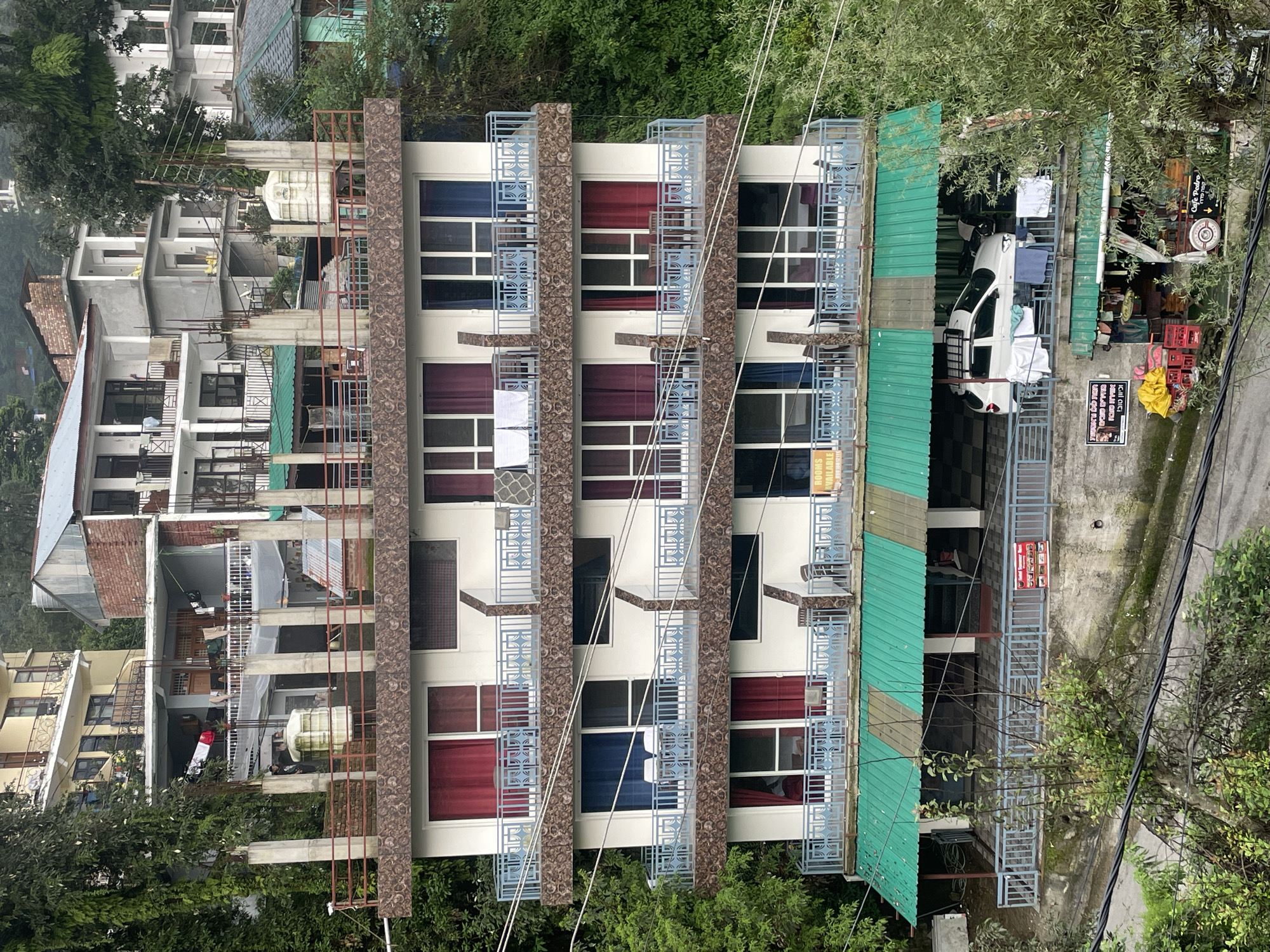 The image shows a multi-story building in Bhagsu, India, with a hotel or guesthouse facade. The building has a brown, textured facade and blue balconies. It features multiple levels with rooms, some of which have red and blue curtains visible through the windows. There are balconies with chairs and tables. A green metal awning covers the ground floor, beneath which a white car is parked. Clothes are hanging out to dry on a railing in front of the car. Below the awning, a small shop with goods displayed is visible. A sign with Hebrew writing and an image of food is posted on the shop. The building is surrounded by lush green trees and foliage. The image appears to have been taken from an elevated position looking down at the building.