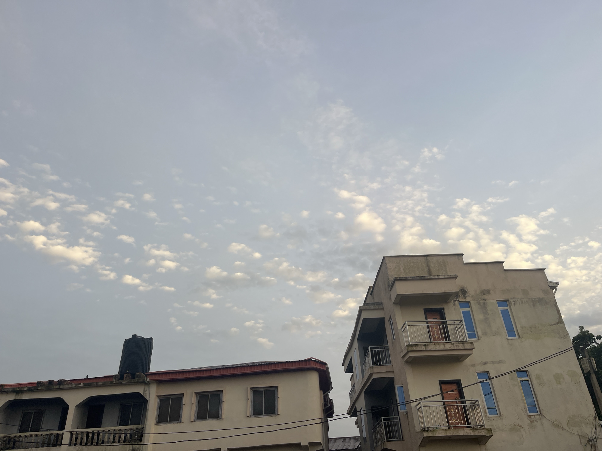 The image depicts a view of buildings and the sky in Jos, Nigeria. Two multi-story buildings are visible, one on the left and another on the right. The building on the left is lighter in color and appears to have a water tank on its roof. The building on the right is taller and has a more modern design, with multiple balconies and windows. The sky is filled with scattered clouds, suggesting it might be early morning or late afternoon. The overall scene is a snapshot of urban life in Jos, showcasing the architecture and the natural elements of the environment.