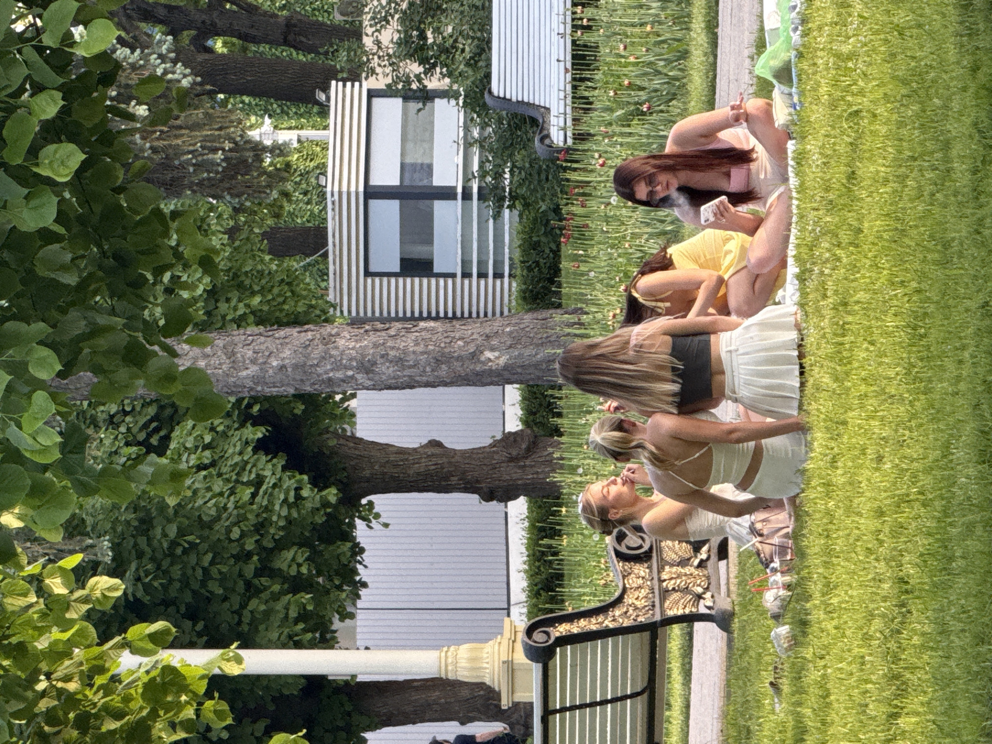 Four young women are gathered on a patch of grass near a park bench. One woman is lying down, another is bent over, and the other two are sitting up. The background features a tree, grass, and a building with white siding. The location is Moscow, Russia.