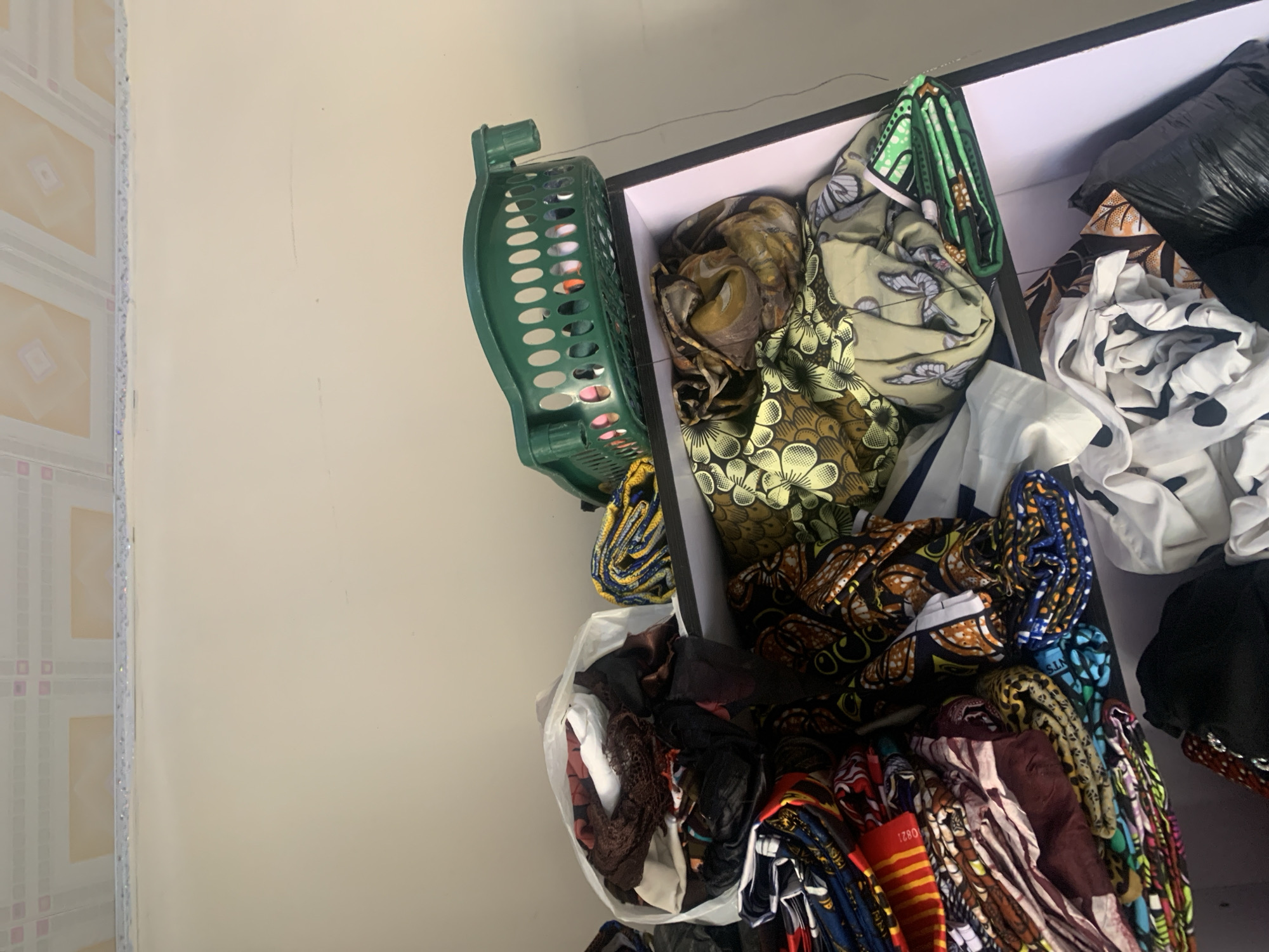 The image depicts a shelf unit filled with folded fabrics and a green plastic basket. The shelf unit appears to be located indoors, possibly in a storage area or a shop. The fabrics are multicolored and feature various patterns, including floral designs and geometric prints. Some fabrics are bundled in a white plastic bag. The green basket, placed on top of the shelf, contains additional folded fabrics. The background shows a textured ceiling and a plain wall, suggesting an interior setting. There are no people visible in the image, and the scene is static.