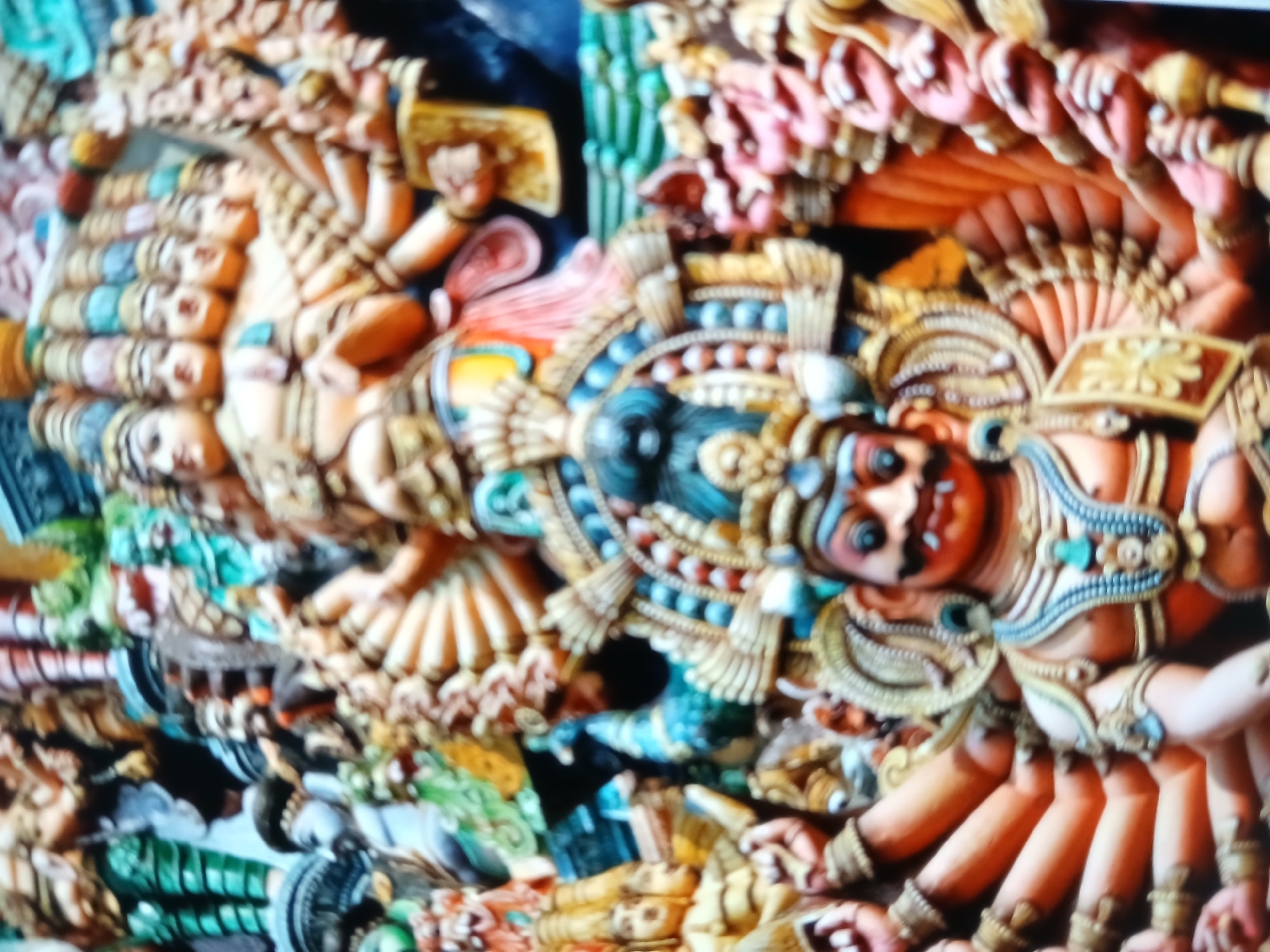 Close-up photograph of a brightly colored Hindu temple carving. The carving depicts a central, stern-faced figure with multiple arms, adorned with ornate jewelry and headgear. To the left, a series of heads are stacked vertically, connected by a similarly ornate band. The backdrop is a mix of green, blue, and black, partially obscured by the figures. The carving is highly detailed with a variety of colors and intricate patterns. This image is likely from a temple in Bengaluru, India.