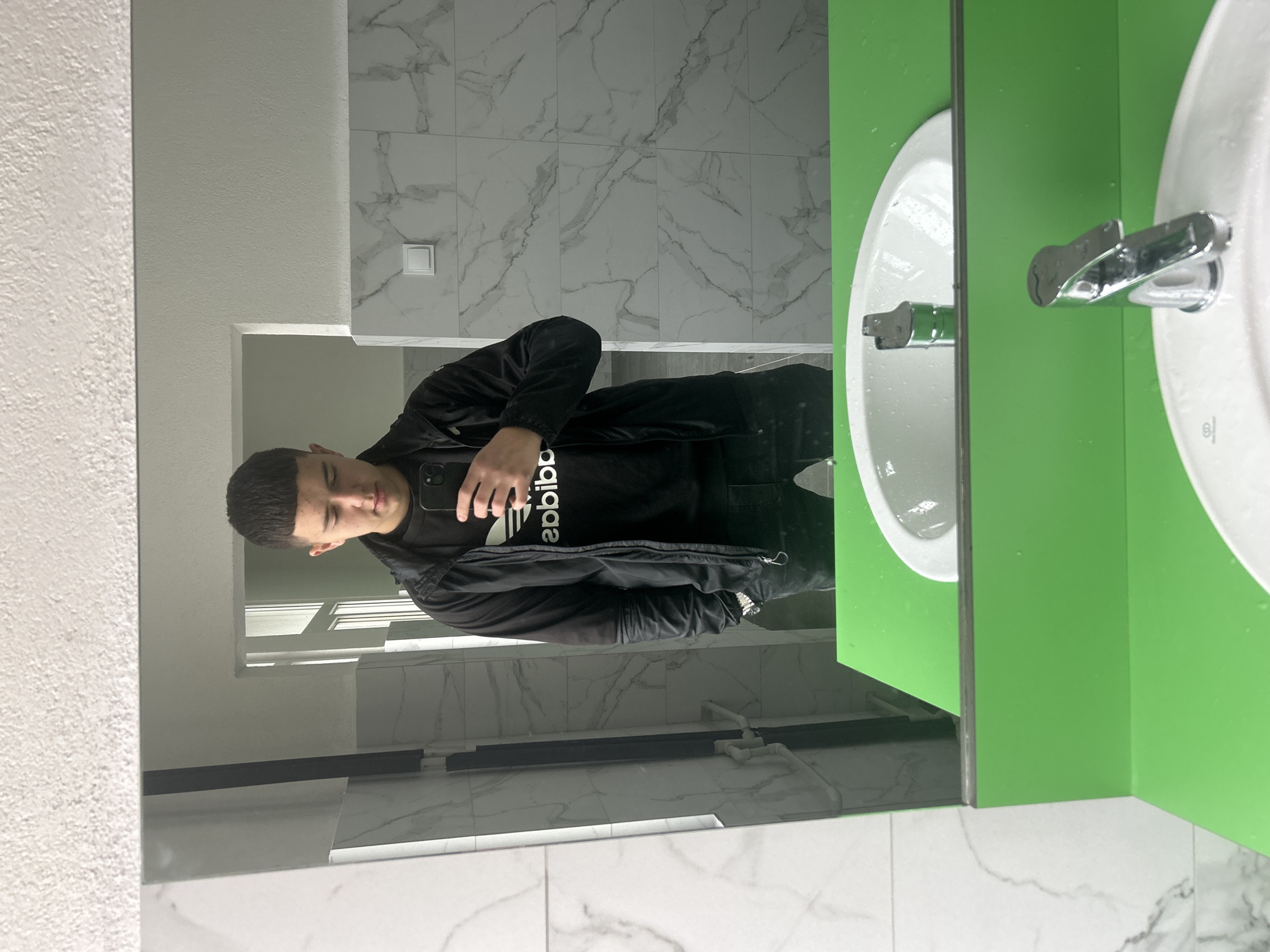 A young man is taking a selfie in a bathroom mirror. The bathroom has white marble tiles on the walls and floor. A sink with a green counter is visible to the right of the mirror. The man is wearing a black Adidas tracksuit. The location is Zlatograd, Bulgaria.