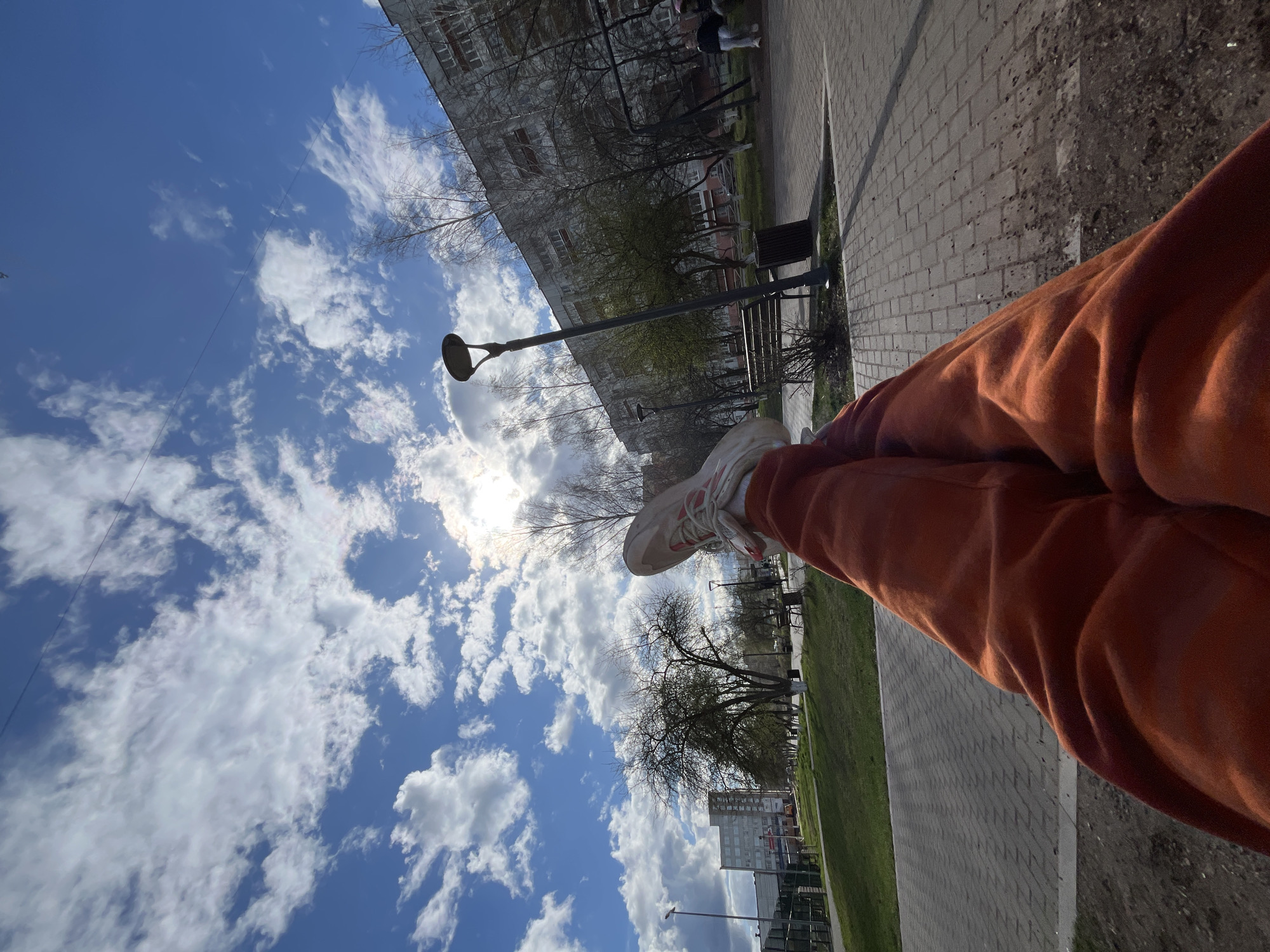 The image, taken from a low angle, depicts a person relaxing outdoors in what appears to be an urban or suburban park setting in Chaikovsky, Russia. The person's lower body is visible in the foreground, wearing orange sweatpants and white and orange sneakers. Their legs are crossed at the ankles and elevated, suggesting they are seated on a bench or similar surface.
The scene features a paved walkway made of grey interlocking bricks, running alongside a strip of green grass. Beyond the path, several deciduous trees stand, some with bare branches characteristic of early spring, and others beginning to show new foliage. Dark park benches and street lamps are positioned along the path. In the background, multi-story residential buildings with multiple windows are visible, hinting at a populated area.
The sky dominates the upper portion of the image, a vibrant blue with numerous fluffy white cumulus clouds. The sun, partially obscured by clouds, is bright and visible, casting strong daylight across the scene. The weather appears clear and sunny. No discernible text is visible on any of the objects or buildings. The overall impression is one of peaceful outdoor relaxation on a bright, early spring day.