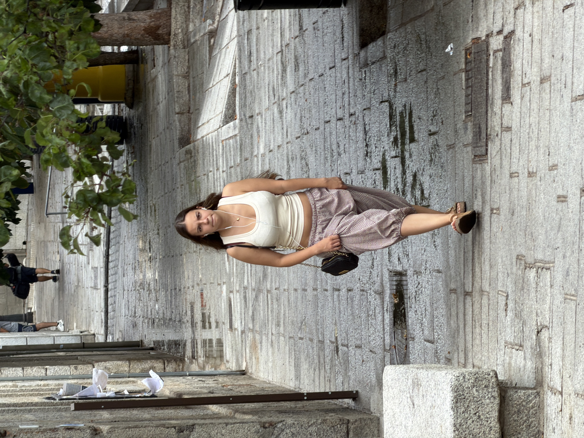 A young woman with long, dark hair and earphones in her ears walks down a sloped, paved street in San Lorenzo de El Escorial, Spain. She is wearing a white tank top, patterned cropped pants, and sandals. A black purse with a chain strap is slung over her shoulder. The street is made of stone pavers, and there are puddles of water on the ground, suggesting it may have recently rained. Buildings with stone walls line one side of the street. In the background, a man carrying a large bag walks away from the camera, and a yellow cylindrical object and trees are visible. The lighting suggests it is daytime.