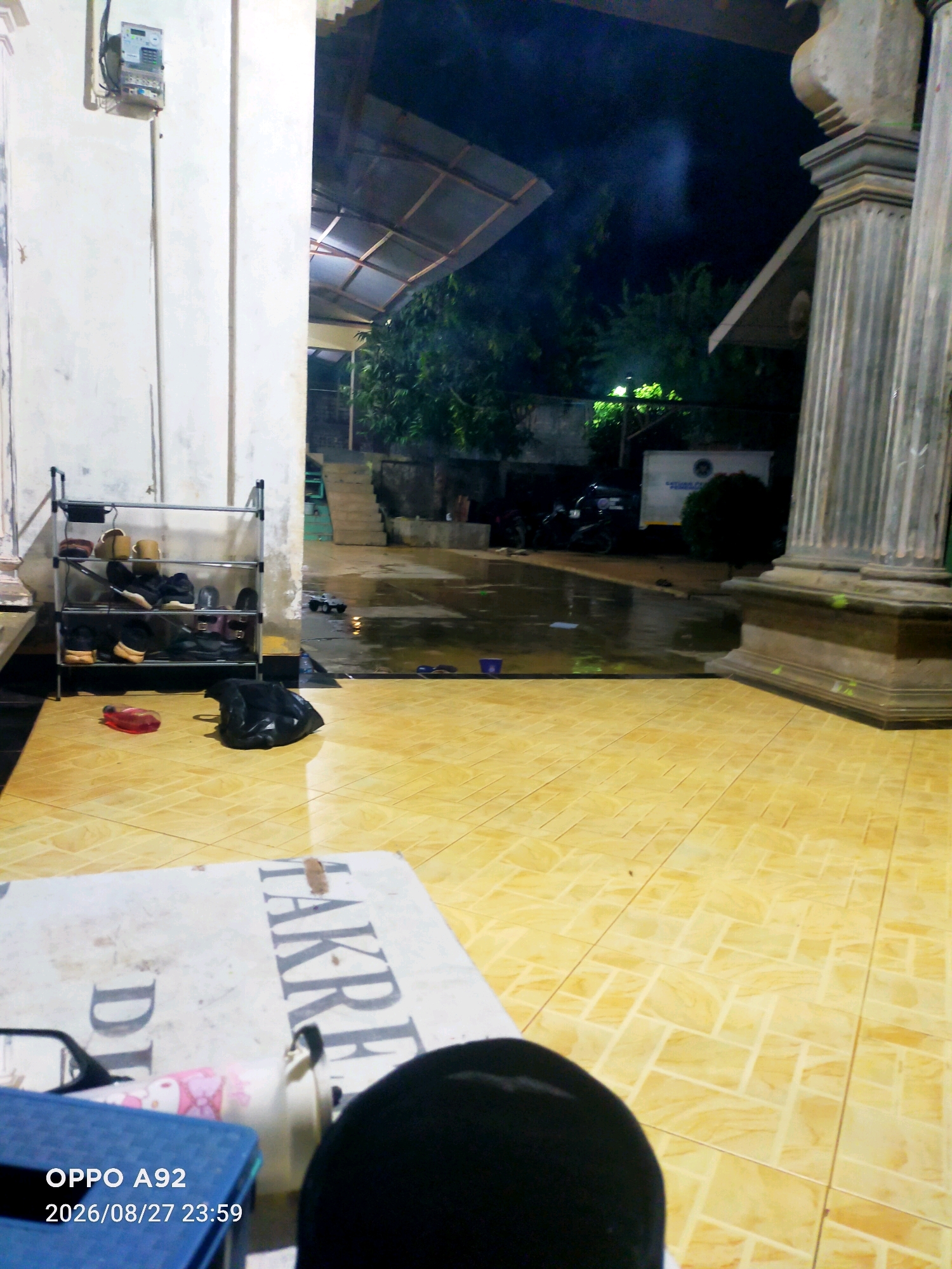 An outdoor nighttime scene in the city of Suka Maju, Indonesia, shows a tiled floor leading to a wet, outdoor area with a covered roof. To the left, a white wall features an electrical meter. In the foreground, a white surface with dark lettering, possibly a table or mat, is visible. A shoe rack with several pairs of shoes is also present on the left. On the wet ground in the mid-ground, a toy car and some scattered items can be seen. In the background, trees and a building with a white front are visible. The lighting suggests a residential or communal outdoor space.