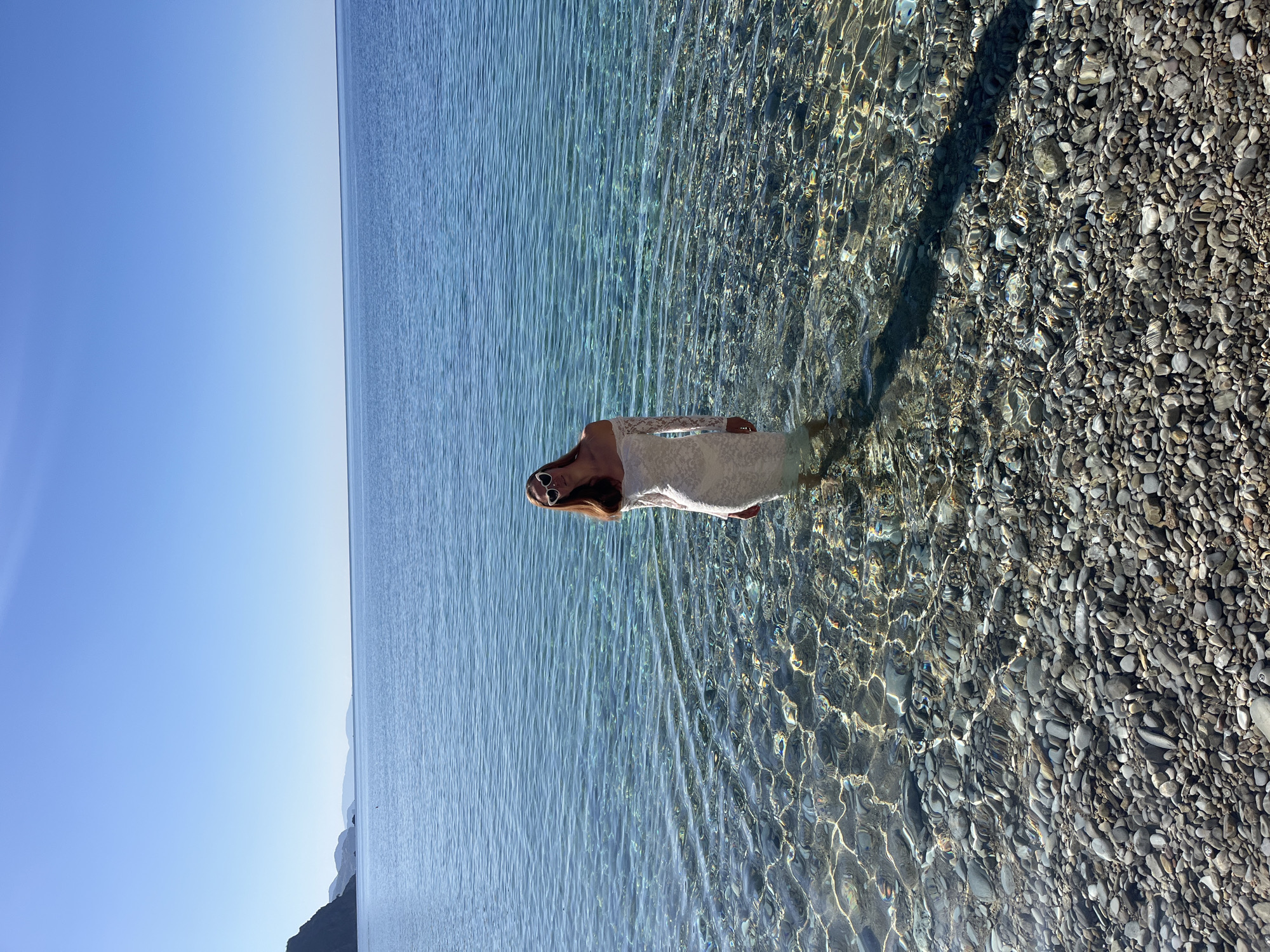 A medium-shot photograph captures a woman standing in clear, shallow water along a rocky shoreline, likely in or near Budva, Montenegro, as implied by the landscape. The water is predominantly clear with visible reflections and refractions on the rocky seabed. The woman is positioned approximately in the center of the frame. The sky is a clear blue, suggesting a bright day. The horizon line is visible in the upper portion of the image. The shoreline is covered in pebbles and rocks, and some mountainous land formations are visible in the distance.
