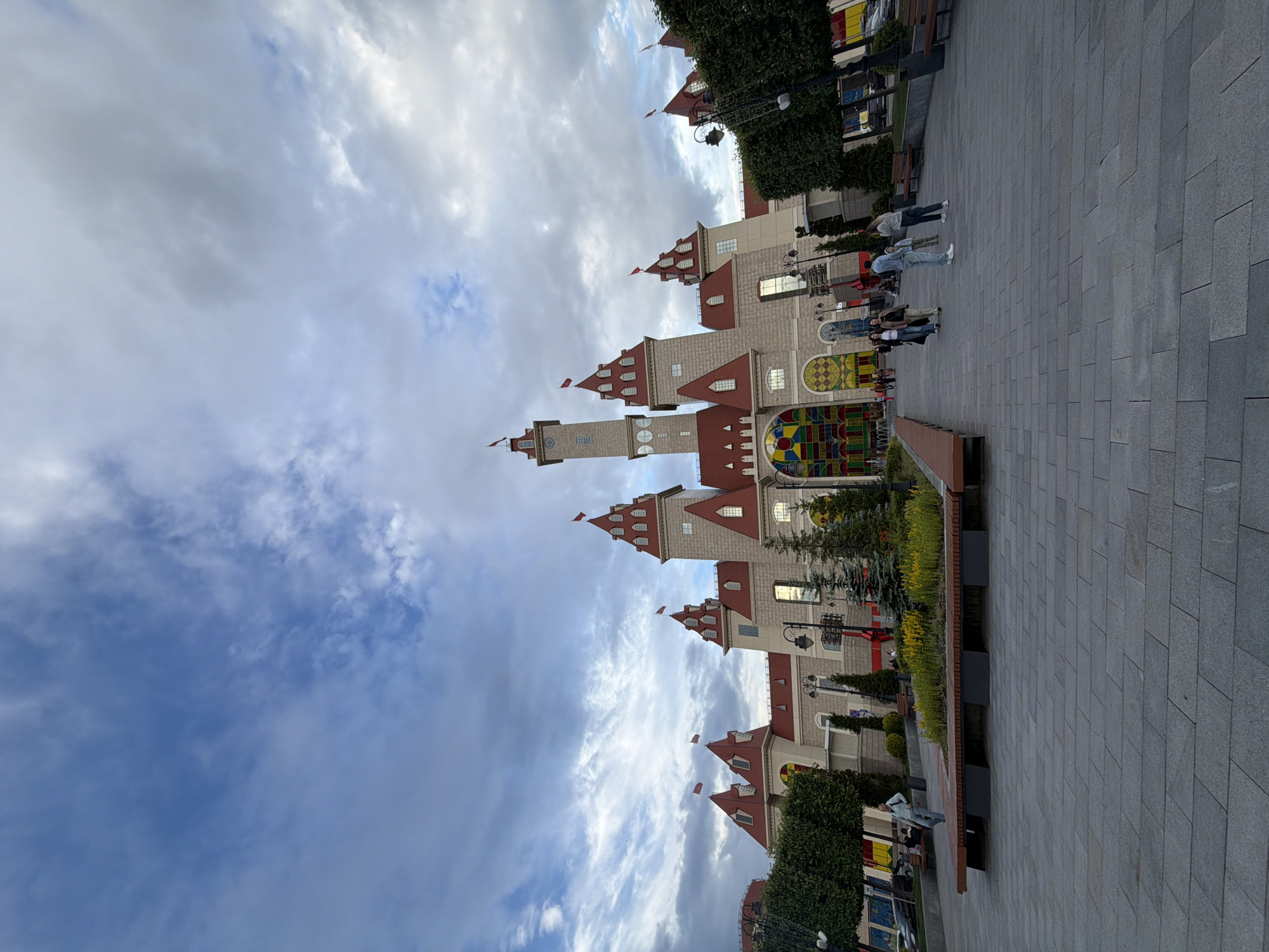 The image shows a wide-angle shot of a castle-like building with red-tiled roofs and multiple towers under a cloudy sky. In the foreground, a paved plaza with benches and landscaping is visible. Several people are walking and standing in the plaza. The building appears to be an amusement park or entertainment venue, with a large stained-glass window feature at its entrance. The setting is an urban environment, with trees and streetlights lining the plaza.
