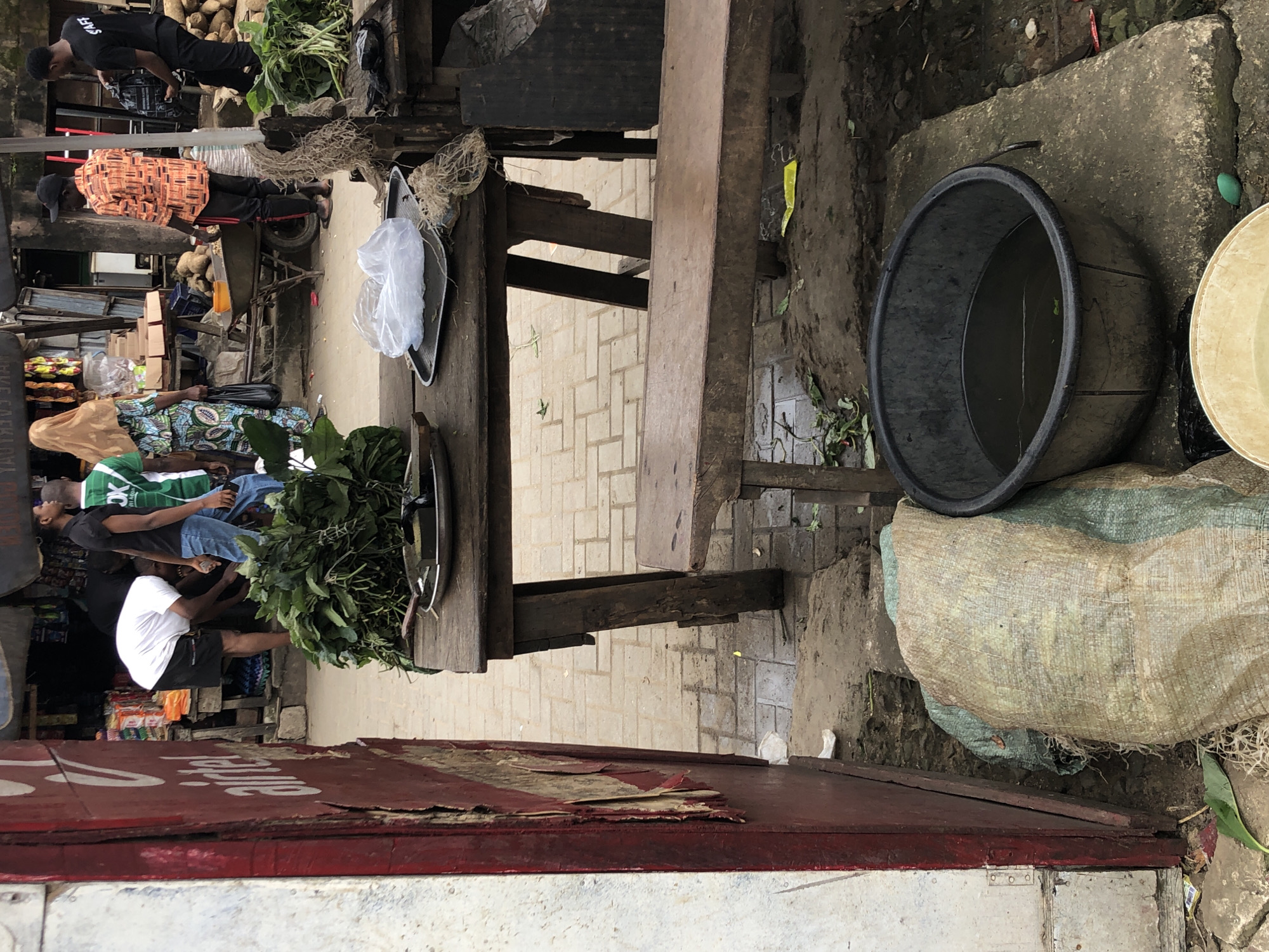 This image depicts a street scene in Ikeja, Nigeria, likely a market area. In the foreground, a large, dark plastic basin filled with murky water sits on a concrete slab. To the left, a large, burlap sack with green sacking is visible. Behind this, a weathered red structure, possibly part of a stall or building, features peeling paint and exposed wood.
In the mid-ground, a rustic wooden table displays a bunch of leafy green vegetables on a metal tray, alongside a plastic bag containing white material and some dried fibrous material. Another wooden bench is positioned in front of the table.
Further back, several individuals are present. One person in a white t-shirt and black shorts is bent over, interacting with someone off-camera. Another person wears a green t-shirt and jeans, and a third person is dressed in an orange patterned shirt and black trousers. A woman in a patterned dress and headscarf is also visible, holding a black plastic bag. In the background, various market stalls are set up, with goods and products displayed. The ground is paved with light-colored bricks and appears wet in places, suggesting recent rain or cleaning. The overall environment suggests a busy, outdoor marketplace.