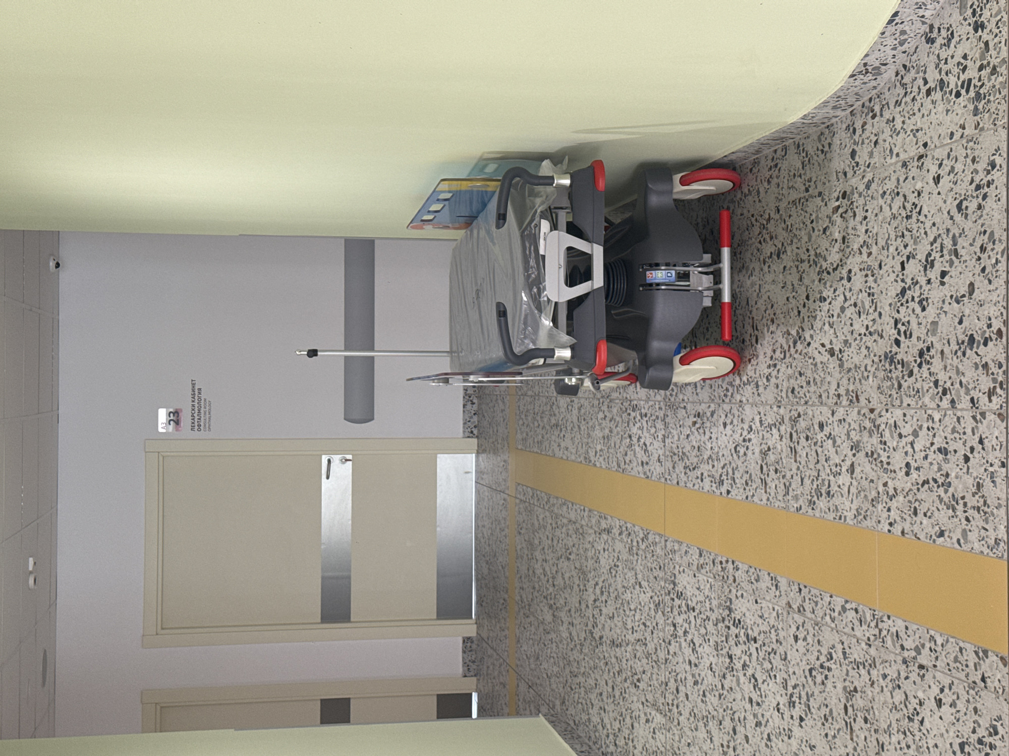 The image depicts a hospital corridor in Burgas, Bulgaria. A light-colored door is visible on the left, with a sign above it indicating "A3 23 ЛЕКАРСКИ КАБИНЕТ ОФТАЛМОЛОГИЯ" and "CONSULTING ROOM OPHTHALMOLOGY". In the center of the corridor, a stretcher covered in plastic is parked, with red and white wheels. The floor is tiled with a speckled pattern and features yellow tactile paving. The walls are painted light grey and a pale green. A security camera is mounted on the grey wall in the upper portion of the image. The scene is devoid of any individuals.