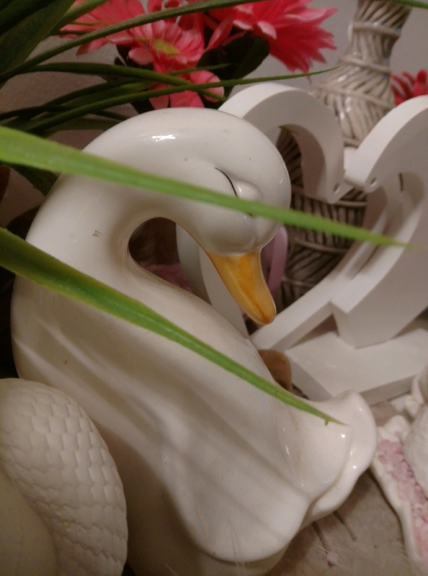 This is a close-up, eye-level shot of a white ceramic swan figurine, positioned prominently in the foreground. The swan is facing to the right, with its head tilted down and its beak, a bright orange, resting on its body. Its eye is a simple black line. Behind the swan, a decorative arrangement of pink artificial flowers and green foliage is visible, with some of the greenery overlapping the swan. Further in the background, a white, abstract decorative piece with curved shapes and perforations is partially visible. The overall impression is that of a decorative item or a display piece. There is no indication of the city of Wedel, Germany, or any specific location within it.